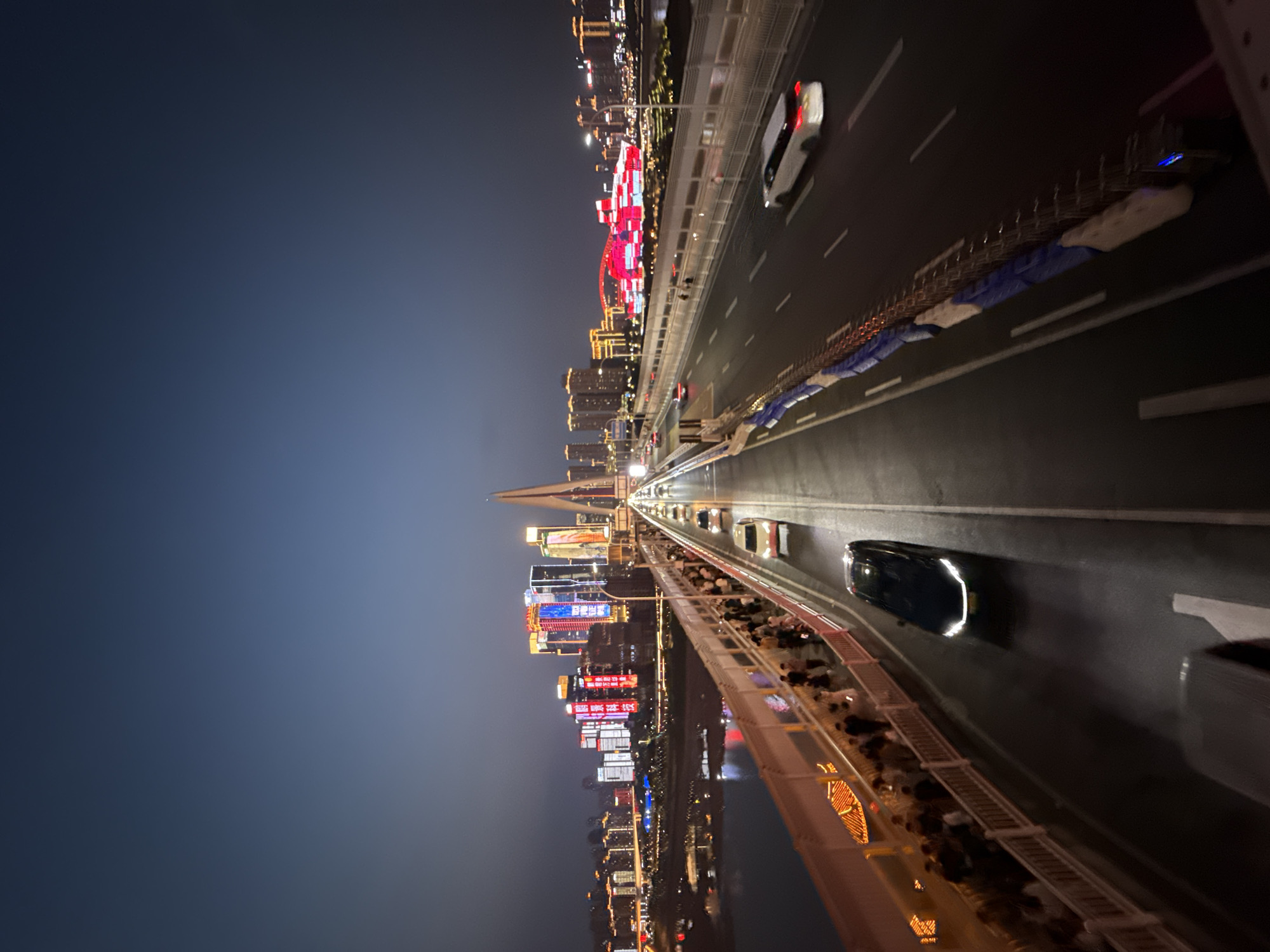 Here is a description of the image:
The image shows a bridge at night in Yuzhong District, Chongqing, China. The bridge is lit up with lights and has cars driving on it. The city skyline is visible in the background, also illuminated with lights. A pedestrian walkway runs along the bridge, and people are walking on it.