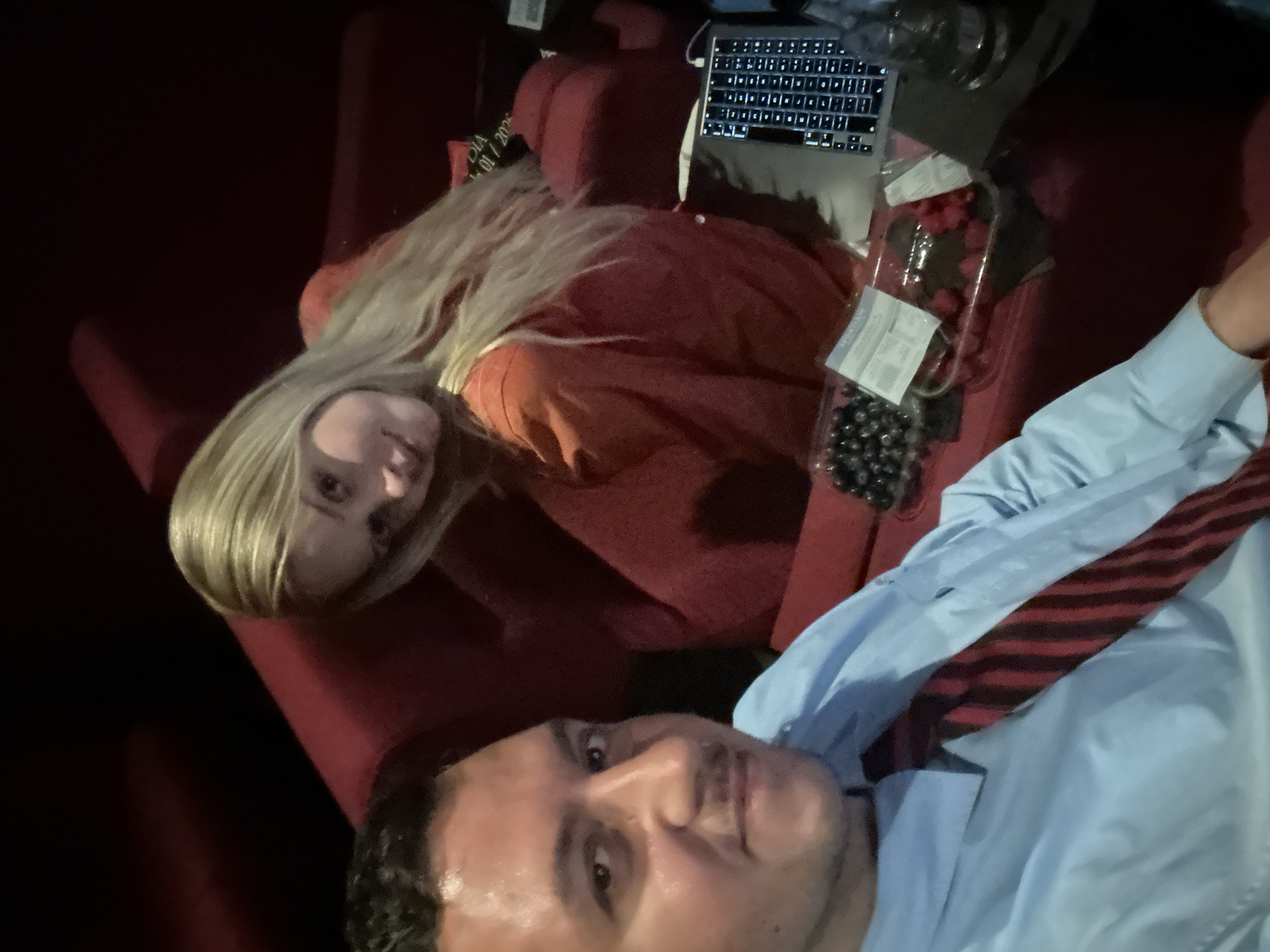 A selfie, taken indoors, shows a man and a woman on a red couch. The man is in the foreground, wearing a light blue dress shirt and a red and black striped tie. The woman, with long blonde hair and wearing an orange shirt, is behind him and to the left. Between them on the couch are a container of raspberries and a container of blueberries. A laptop is open behind the woman. The location is unknown, it cannot be forensically determined that this picture was taken in London, United Kingdom.