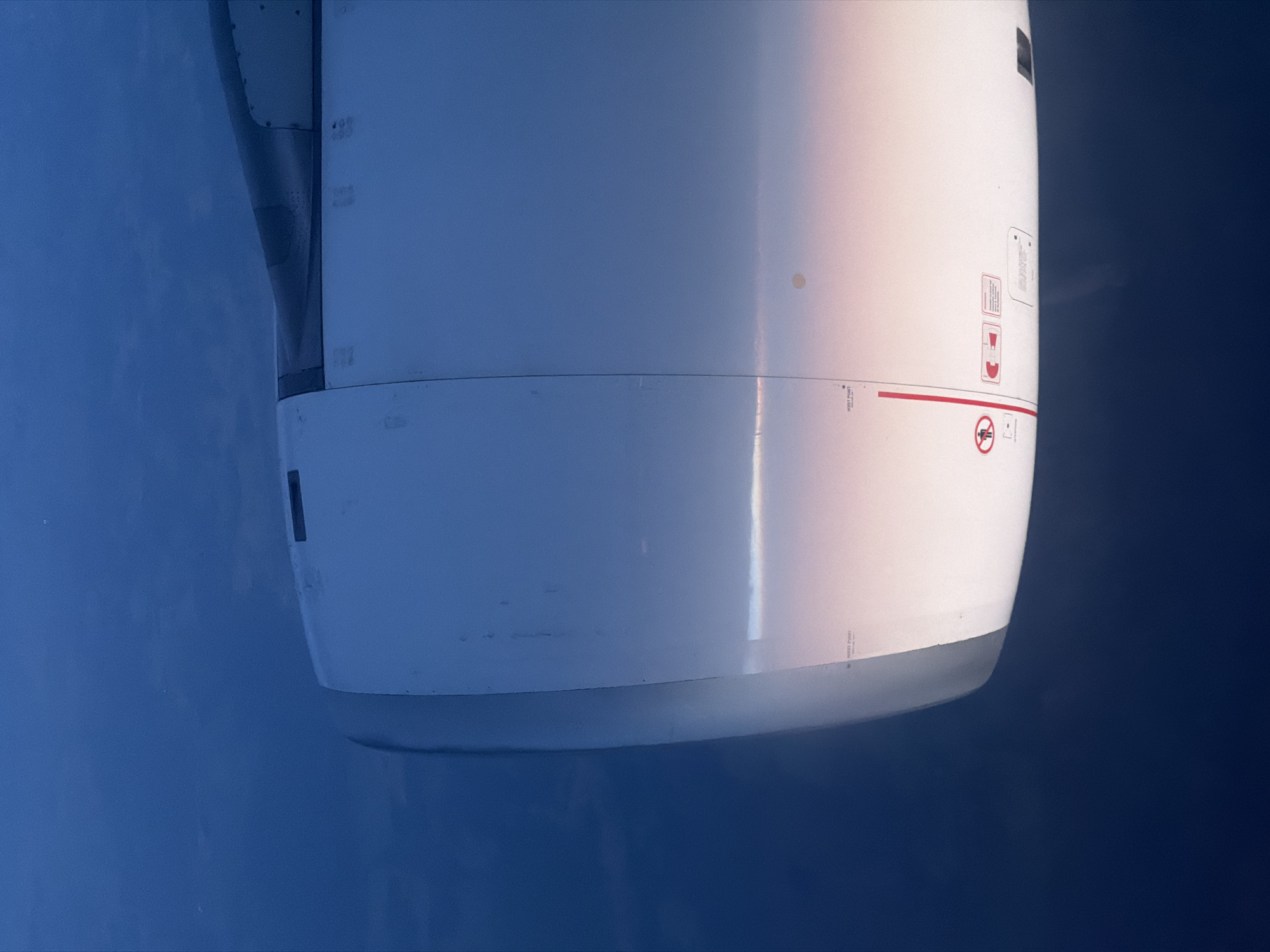 An airplane engine is visible in the frame, positioned in front of a blue sky. The engine is predominantly white with some grey areas, and it features various labels, markings, and a red line. Part of the wing structure is also visible. The scene appears to be taken during flight, with the focus on the engine against the sky. The location is not specified.