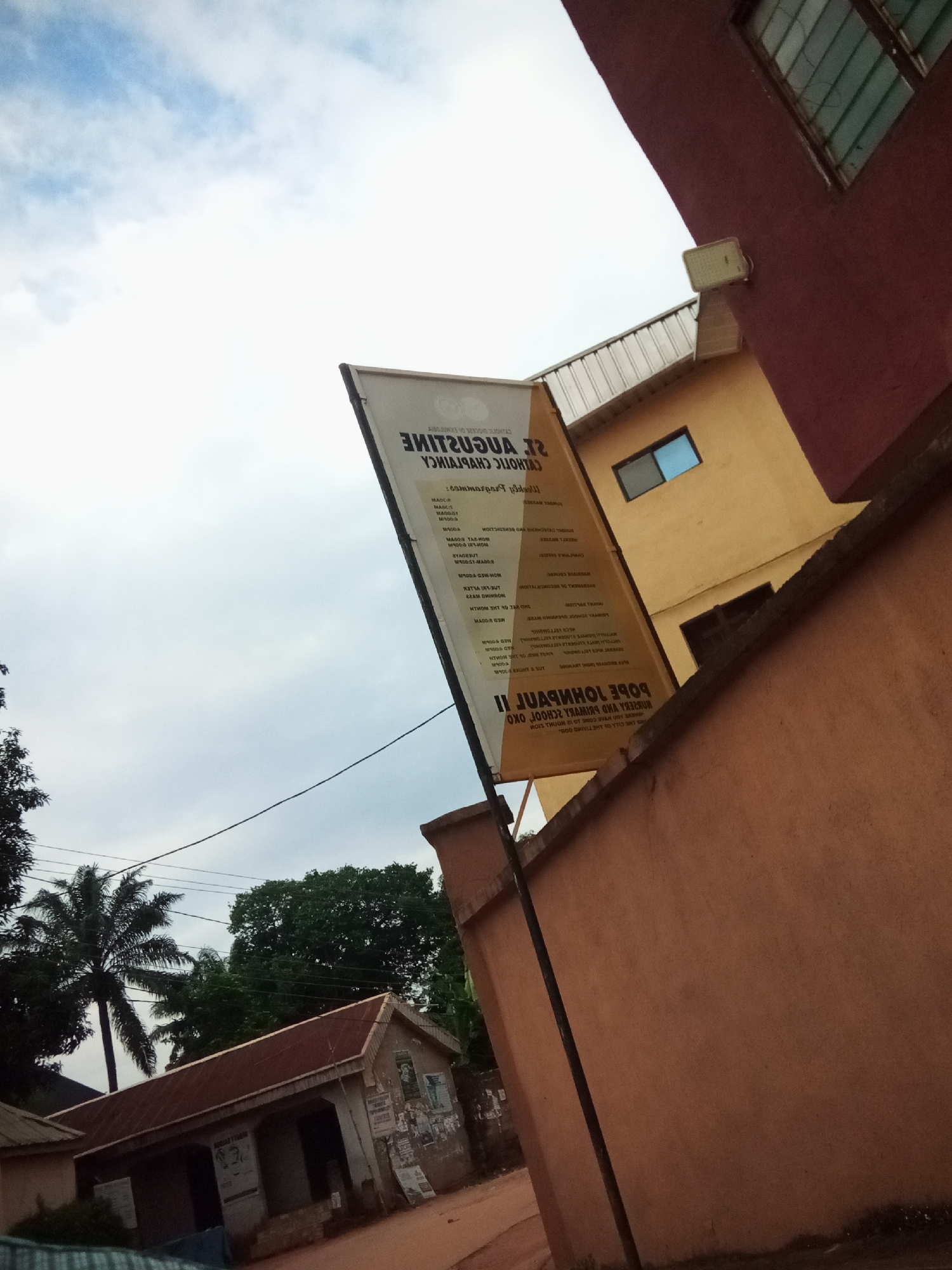 The image depicts an exterior scene in Oko Town, Nigeria. A large, rectangular banner, angled towards the sky, is prominently displayed. The banner is white with black text, some of which appears to be reversed or upside down. Text visible on the banner includes "ST. AUGUSTINE CATHOLIC CHAPLAINCY" and "POPE JOHNPAUL II NURSERY AND PRIMARY SCHOOL OXO."
In the background, two buildings are visible. The building to the right is a multi-story structure painted a deep red, with a window that has green shutters or bars. The building to the left is painted yellow and has a corrugated metal roof. A smaller, light-colored building with a reddish-brown corrugated metal roof is situated in the lower left quadrant of the image. This building appears to be a school or community building, with posters and notices affixed to its exterior walls.
A tall, slender palm tree is visible behind the school building. Dense green foliage from trees is also present in the background, suggesting a tropical or sub-tropical environment. The sky is overcast with light clouds. A thin, dark pole runs vertically up the right side of the image, in front of the red building. Electrical wires are strung across the upper portion of the scene. The ground appears to be unpaved. The overall impression is that of a community or religious institution in a town setting.