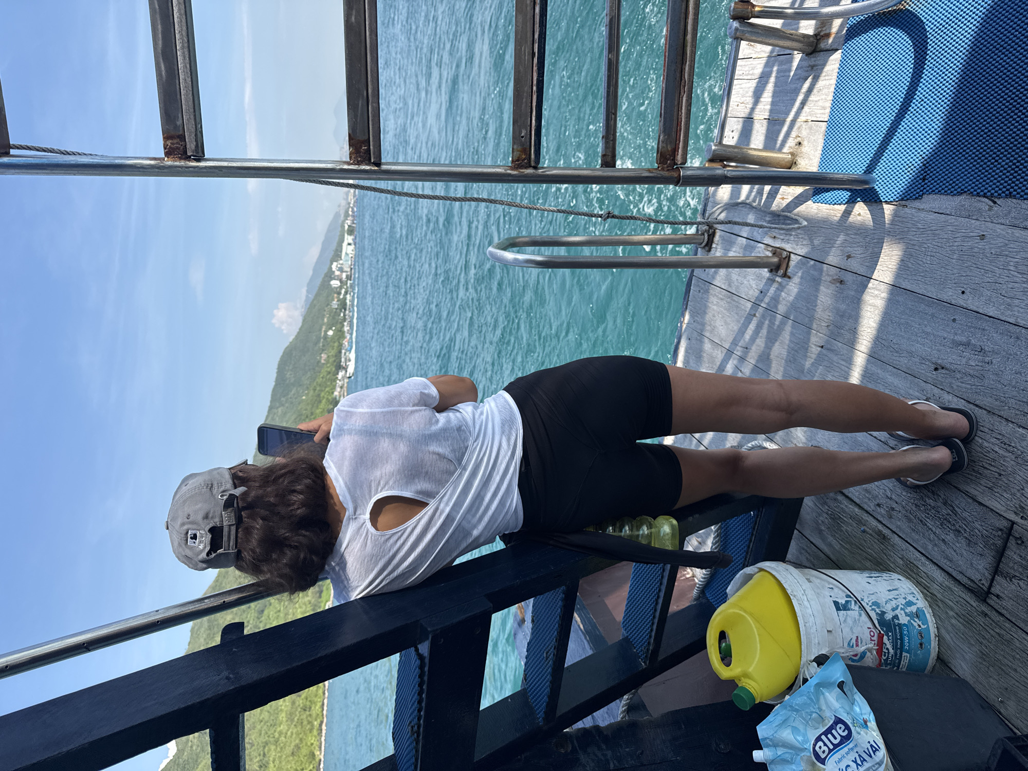 A woman wearing a grey baseball cap, a white t-shirt with a cut-out in the back, and black shorts stands on the deck of a boat. She is holding a smartphone and appears to be taking a picture. The boat is on the water, with a coastline of green hills and buildings visible in the background. The sea is a clear blue, and the sky is bright and sunny with a few scattered clouds. On the deck, near the woman, are a white bucket with a yellow jug and a blue plastic bag. A metal ladder is attached to the side of the boat. The scene appears to be in the waters off the coast of Nam Nha Trang Ward, Vietnam.