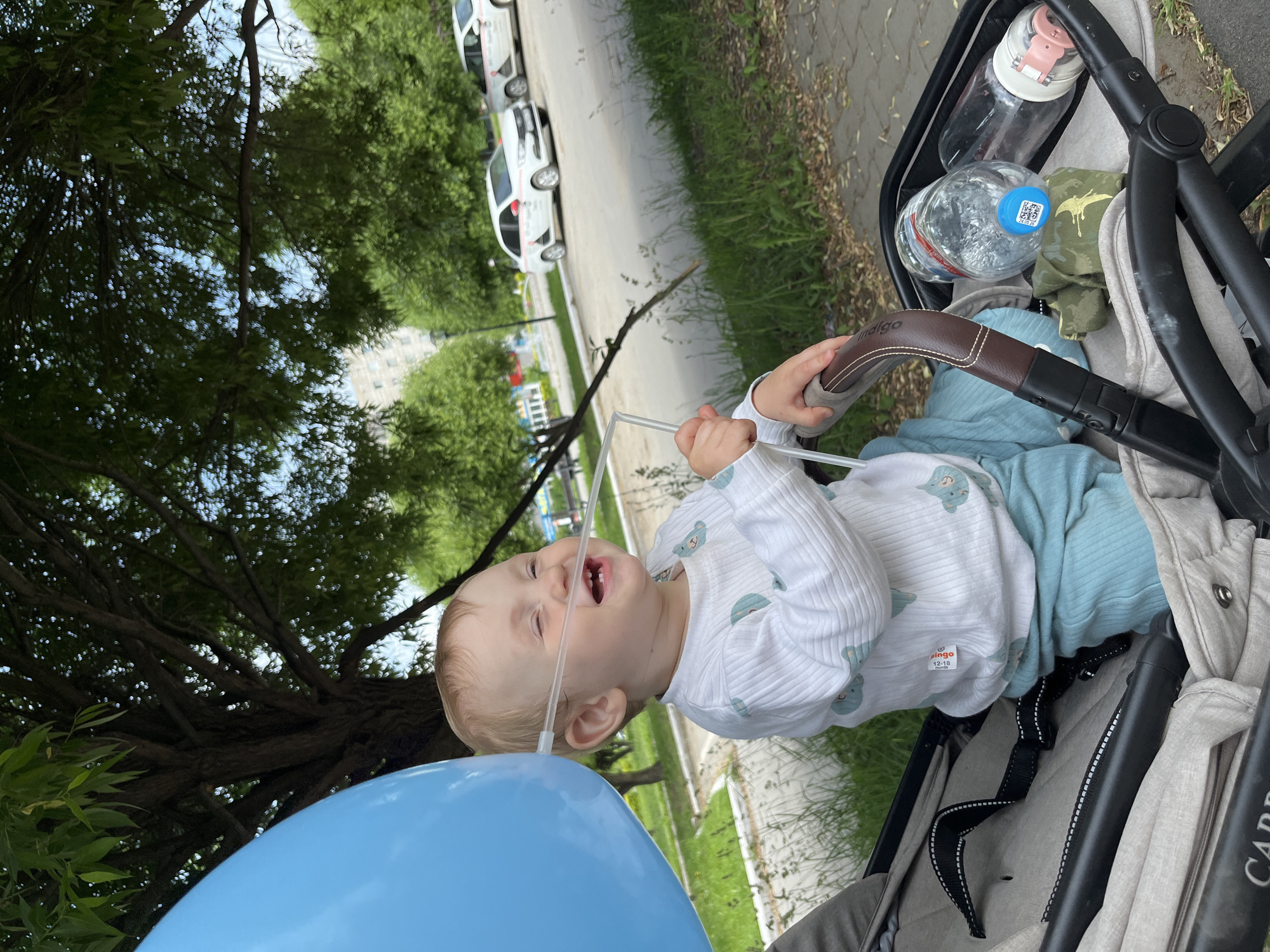 A young Caucasian child with fair hair, wearing a white long-sleeved shirt with a blue teddy bear pattern and light blue pants, sits in a gray stroller. A blue balloon is held to the child's face by a thin tube, and the child is smiling. The stroller is on a paved path next to a grassy area with trees and parked cars in the background. The scene appears to be outdoors in a park or residential area. The weather seems to be clear and sunny. The child appears to be receiving oxygen therapy via nasal cannula. The stroller has a brown handle and a black safety strap. There are two water bottles and a green fabric item in the stroller. The child is looking to the right. In the background, there are buildings and more trees. The location is the city of Chaikovsky, Russia.