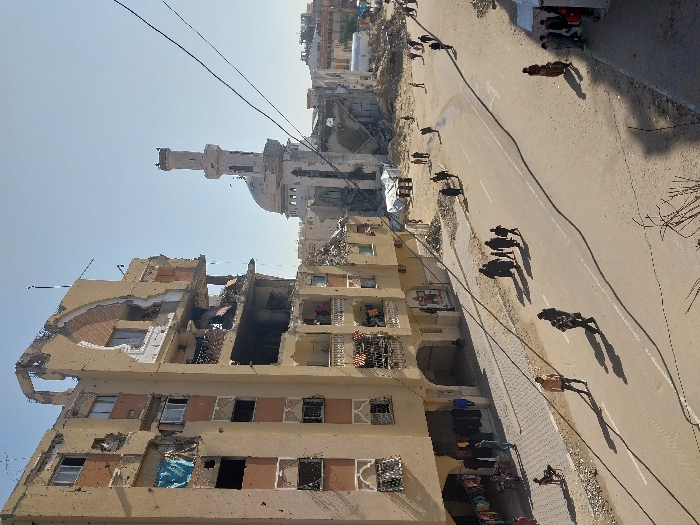 This image captures a street scene in Khan Yunis, Palestinian Territory, under a clear, bright sky. A multi-story building dominating the left side of the frame exhibits extensive structural damage, with large sections of its facade missing, exposing internal rooms and twisted rebar. The building's exterior also shows numerous smaller impact marks. On its ground floor, racks of clothing are displayed outside, and people are visible near these shops. In the mid-ground, a damaged mosque featuring a tall minaret and a dome is partially visible, surrounded by rubble and debris. The paved road running through the center of the image is traversed by numerous pedestrians, including adults and children, moving in various directions. Dirt and sand cover the areas beside the road, where piles of rubble and at least two white tarpaulins are present. Overhead electrical or communication wires crisscross the upper portion of the frame. Long shadows cast by the people and structures indicate strong sunlight.