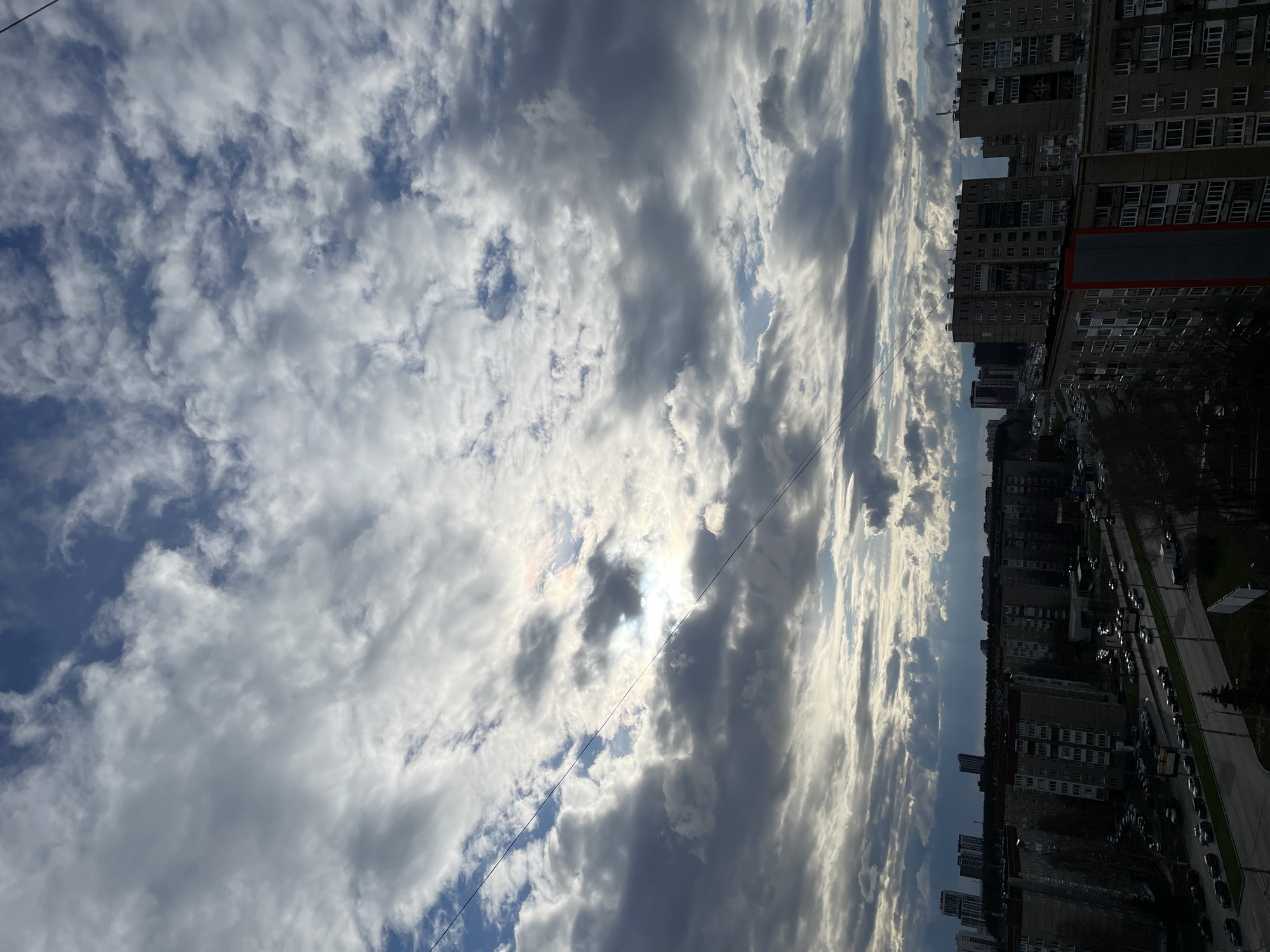 The image depicts an urban landscape under a dramatic, partly cloudy sky, characteristic of Izhevsk, Russia. The sky dominates the upper portion of the frame, filled with a mix of large, dynamic stratocumulus and cumulus clouds, varying in shades of white and grey. Patches of clear blue sky are visible between the clouds. The sun is partially obscured by a central cloud, creating a bright, radiant burst of light that illuminates the surrounding clouds and suggests either late morning or late afternoon.
Below the horizon line, a dense arrangement of multi-story residential buildings forms the cityscape. These buildings, consistent with post-Soviet architecture, feature numerous windows and block-like facades, stretching along a wide street. The street is active with many cars, both parked along the curbs and moving in traffic, though no individual people are distinctly visible. Sparsely scattered trees, appearing bare, indicate a season other than summer, likely late autumn, winter, or early spring. A thin utility cable or power line runs diagonally across the middle of the sky. There is no discernible text visible on any buildings or signs within the frame. The overall scene captures the interplay between an active city environment and a striking, ever-changing sky.
