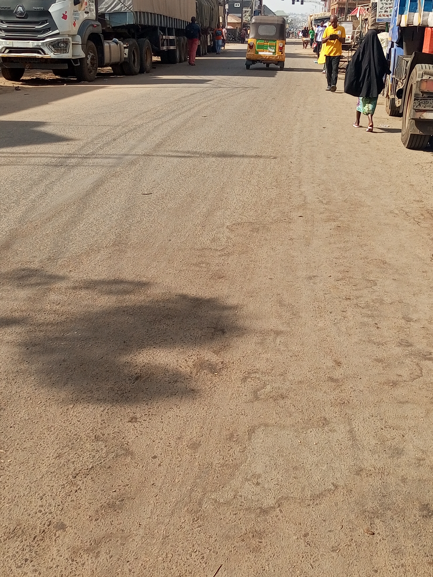 The image shows a street in Maiburiji, Nigeria. The foreground is a beige, asphalt road. In the distance, there are several people walking on the right side of the road. To the left, there are several large trucks. A yellow auto rickshaw is positioned in the center of the road. On the right side, a person wearing a yellow shirt is walking. To the right of the person in yellow, a person dressed in a black hijab is walking. The environment is sunny.