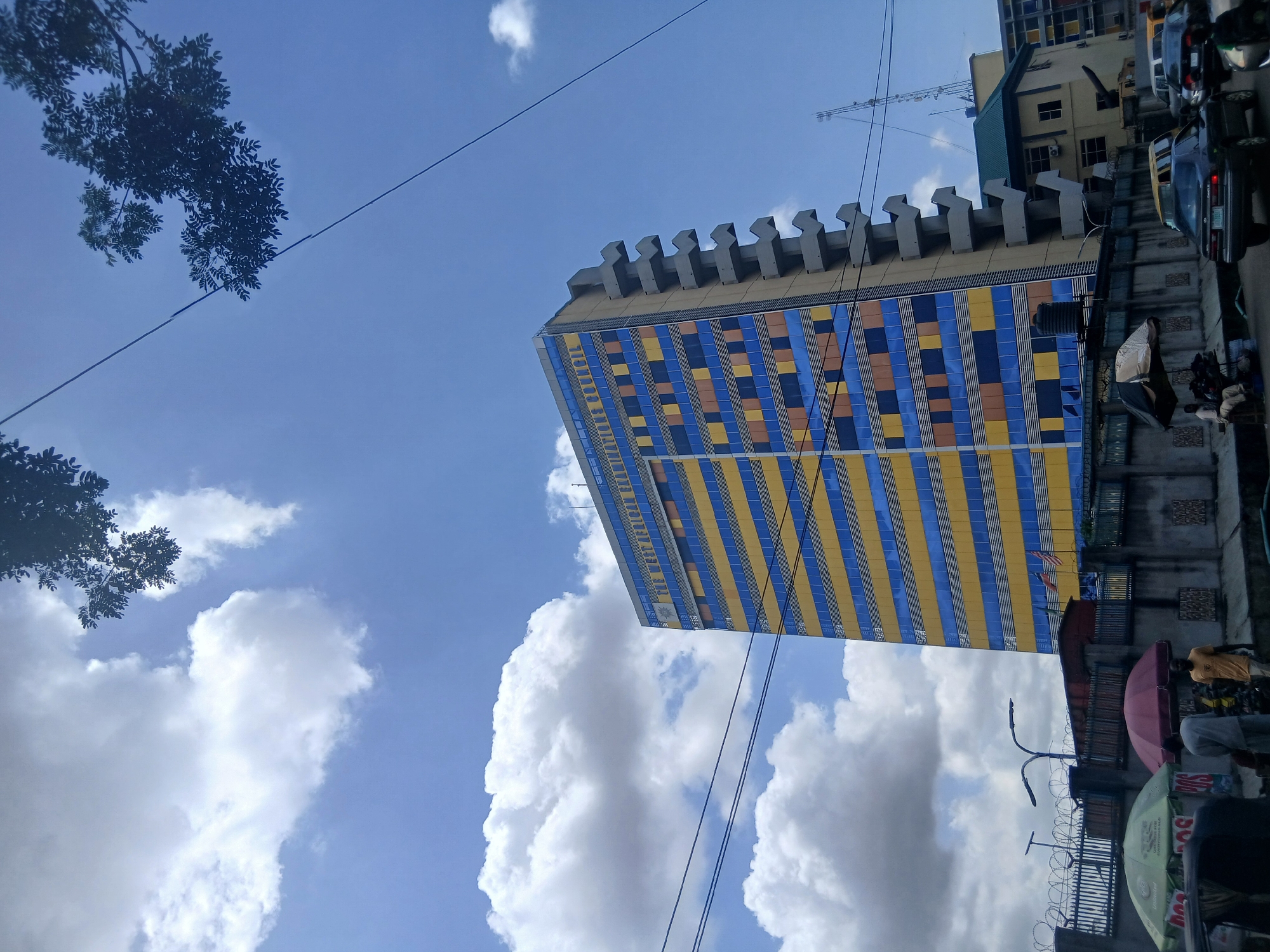 A low-angle, outdoor shot depicts a tall, rectangular building with a distinctive architectural design. The facade features alternating vertical stripes of blue and yellow, with a checkerboard pattern of blue, yellow, and brown squares at the top. A grey, crenellated parapet tops the structure.
The sky is bright blue, dotted with puffy white clouds. In the top left corner, tree branches with dark green foliage are visible. Power lines run across the frame, intersecting with the building and sky.
On the right side of the frame, a portion of another building can be seen, with vehicles parked nearby. Below the building, a cluttered street scene unfolds with figures, possibly vendors and pedestrians, along with umbrellas, carts, and barriers.
This is likely the headquarters building of the Nigerian Petroleum Corporation (NNPC) in Ikoyi, Lagos, Nigeria. The image captures a mix of urban environment, with the architectural landmark set against a backdrop of daily street life.