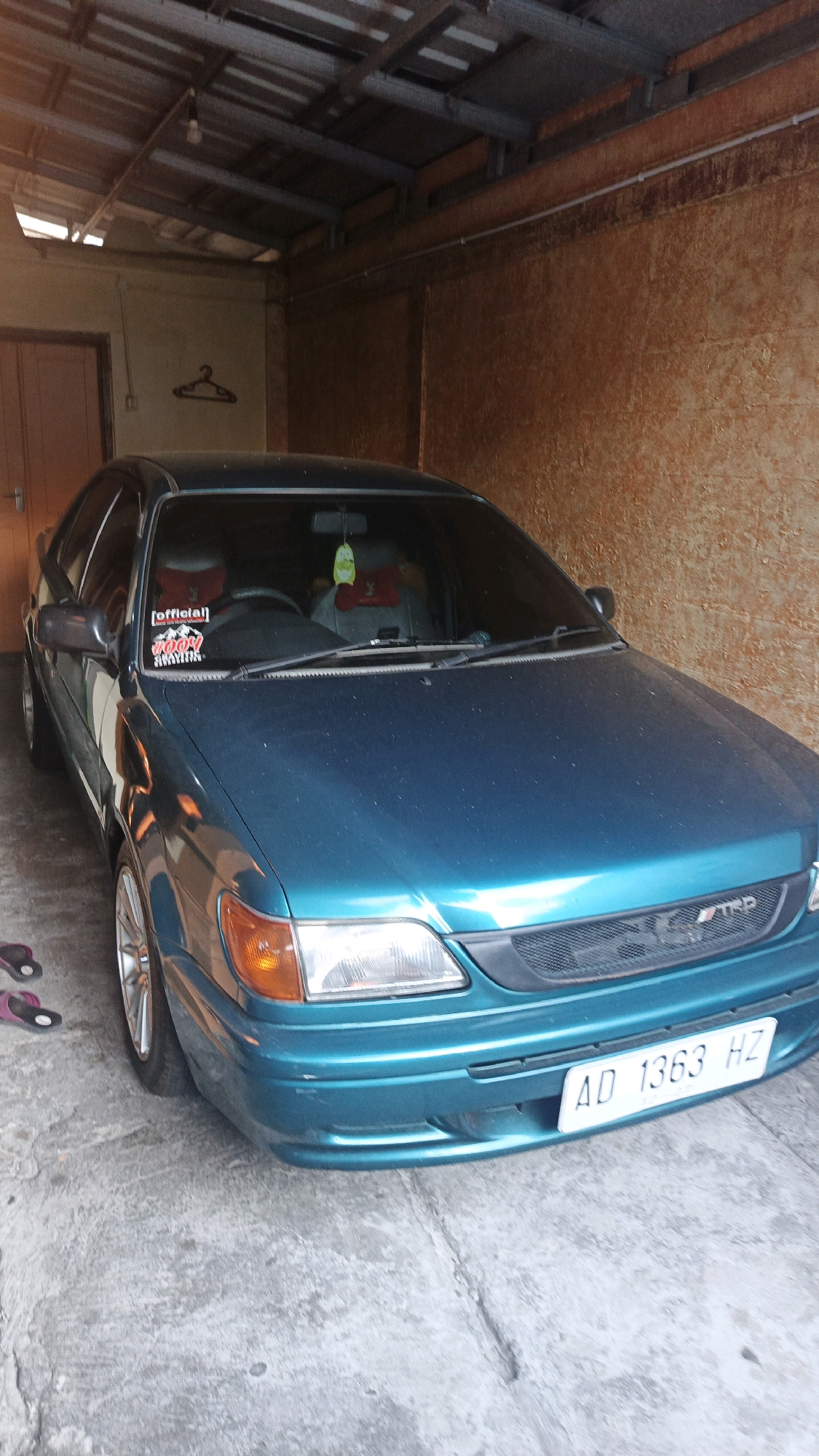 A teal sedan is parked indoors. The car has a black grille with a TRD emblem and a license plate reading "AD 1363 HZ." Stickers are visible on the windshield, one red with "official" and another red and white with "ROOR." Inside the car, red seat covers are on the headrests. The environment appears to be a garage or a covered outdoor space with a corrugated metal roof and textured walls. A wooden door is visible on the left, and a coat hanger is on the wall behind the car. There are no people in the image. The scene is well-lit, suggesting it's daytime. The location is likely in the city of Surakarta, Indonesia, given the license plate format.