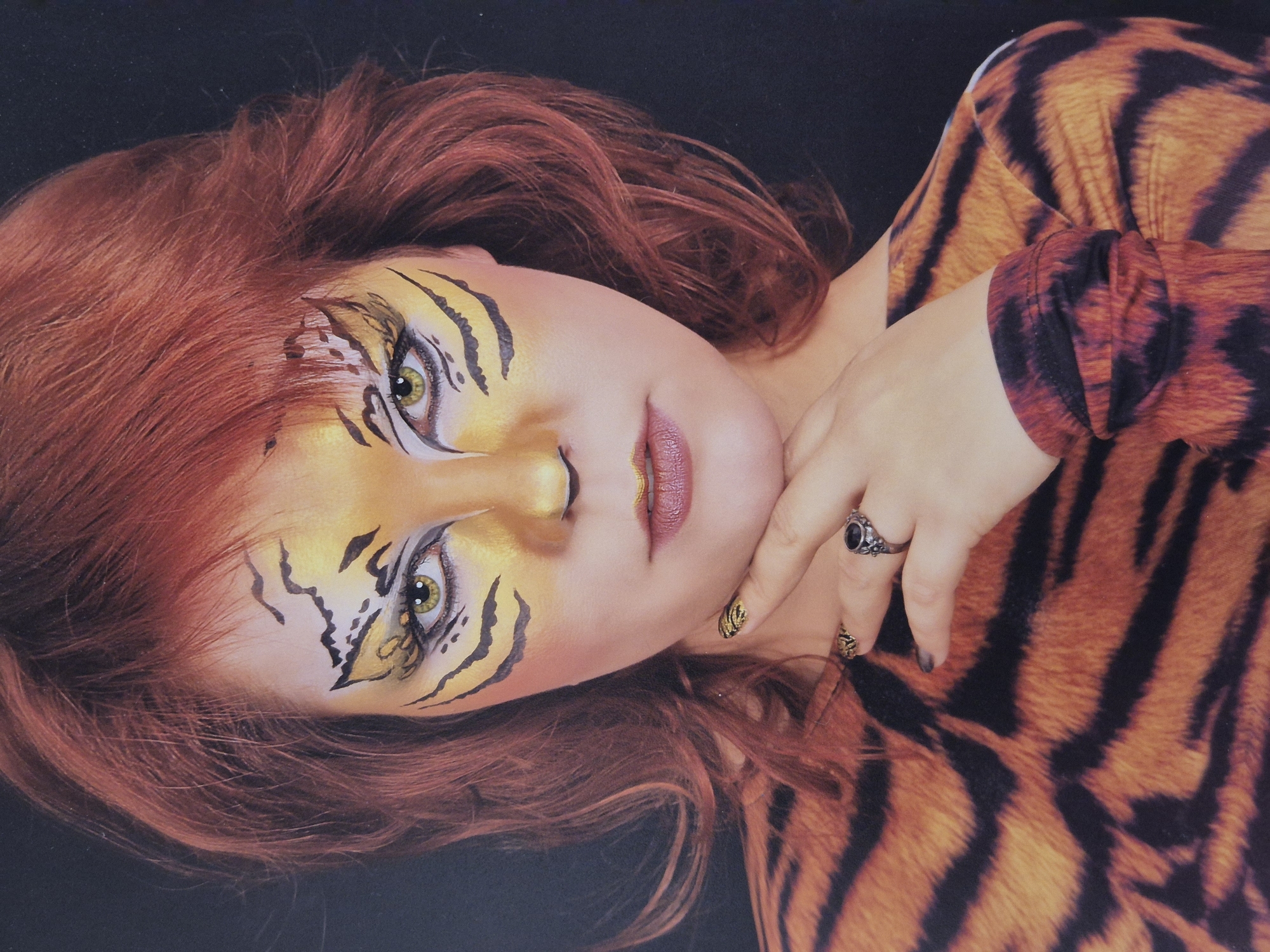 A woman with red hair and tiger-striped makeup is posing against a black background. She is wearing a tiger-striped top and has her hand to her chin. The makeup includes yellow and orange face paint with black stripes around her eyes and on her nose. She is also wearing rings on her fingers. The image appears to be a studio portrait. There is no information about the city of Schwäbisch Gmünd, Germany in the image.