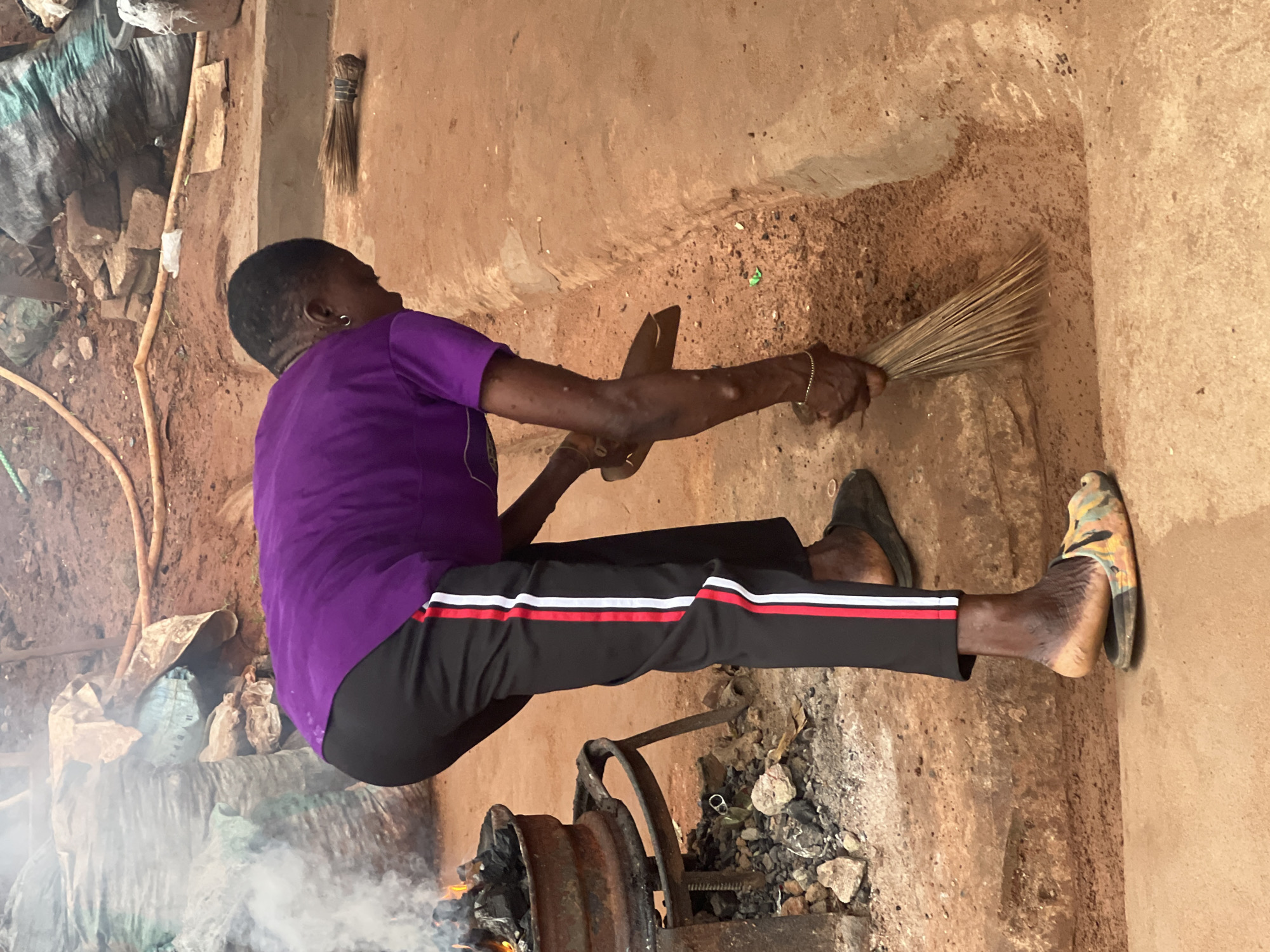A person wearing a purple t-shirt and black pants with red and white stripes is sweeping a dirt path with a broom. The person is bent over, their back to the camera, and their feet are bare except for a pair of colorful, patterned sandals. To the left, there is a metal brazier with a fire burning inside, emitting smoke, and a pile of charcoal. In the background, there are large sacks, some appearing to be filled with charcoal, and a yellow hose. The ground is a reddish-brown dirt. The scene appears to be an outdoor area, possibly a workshop or market, in Nkpor, Nigeria.