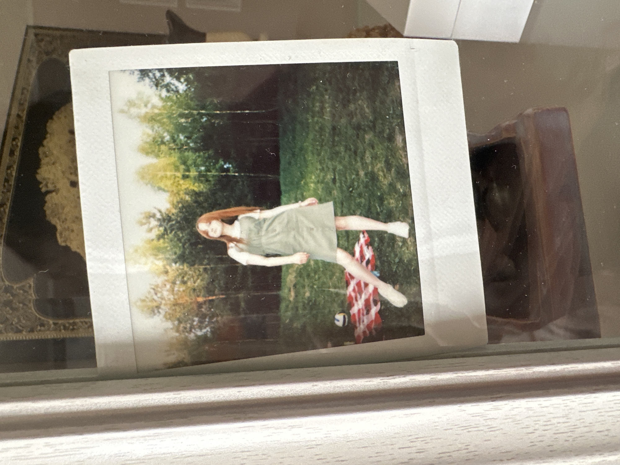 A Polaroid photograph of a woman in a green dress lying horizontally on a grassy area is set against a backdrop of trees. A plaid blanket and a ball are visible on the ground. The photo is placed within a display case or a frame, with other items visible in the background, including a decorative object and a dark-colored rectangular container. The display case is positioned on a light-colored surface. There is no visual information to determine the location in the city of Vitsebsk, Belarus.