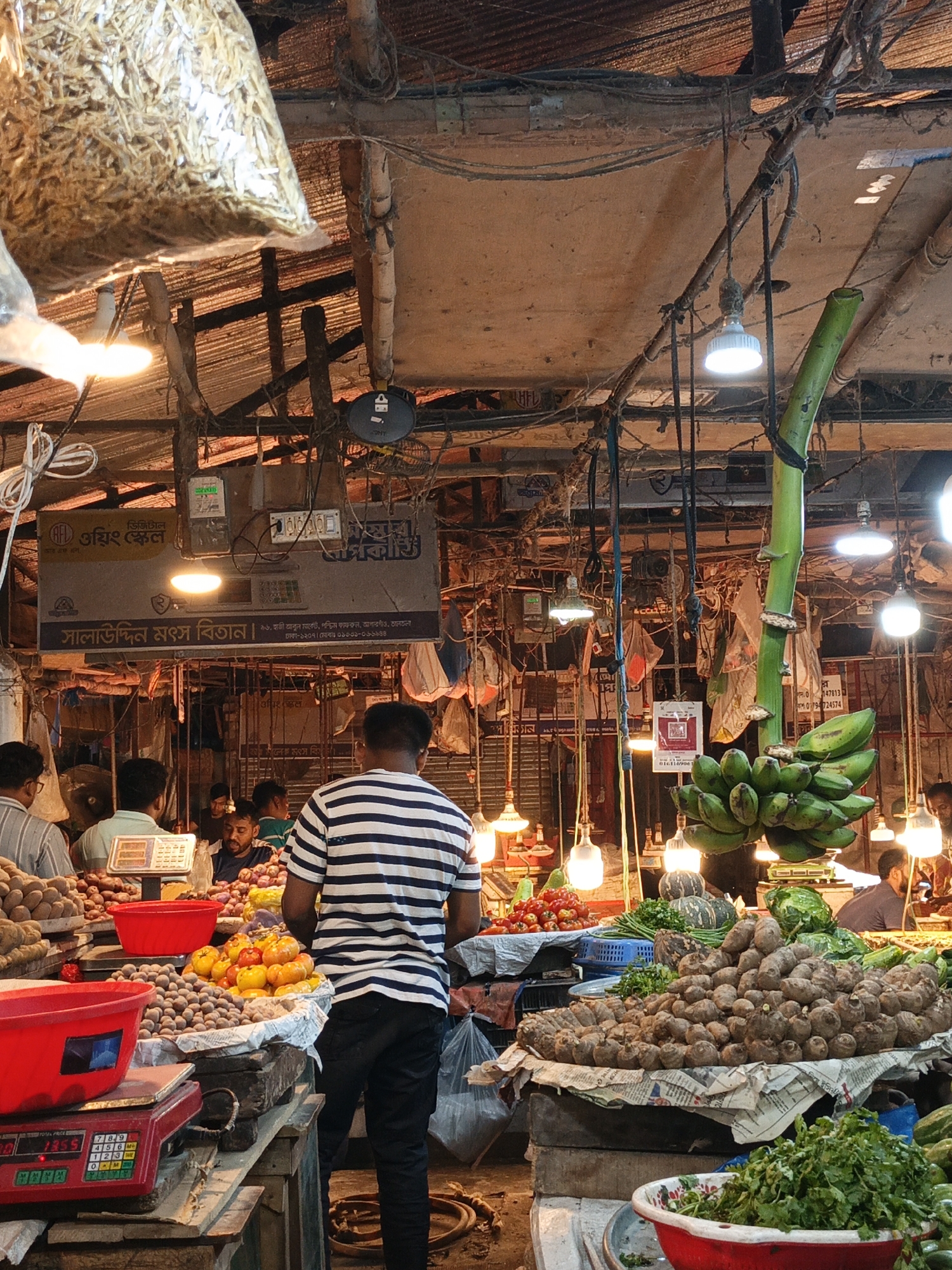 The image depicts a bustling marketplace, likely in Dhaka, Bangladesh, as suggested by the prominent sign "সালাউদ্দিন মৎস বিতান" (Salaluddin Fish Mart) in Bengali, indicating a possible location near Haji Abun Market, Agargaon, Dhaka-1207. The market is crowded with various vendors and customers. In the foreground, there are displays of produce including potatoes, tomatoes, and other vegetables. A person wearing a striped shirt is standing in front of the produce, apparently shopping. A weighing scale is visible in the lower-left corner, presumably for measuring the goods. Overhead, a tangle of wires and exposed infrastructure create a chaotic ceiling. Hanging from the ceiling are several light bulbs illuminating the scene and bunches of bananas tied to a bamboo pole. A bag of dried fish is seen in the upper-left corner. The market setting appears traditional and busy.