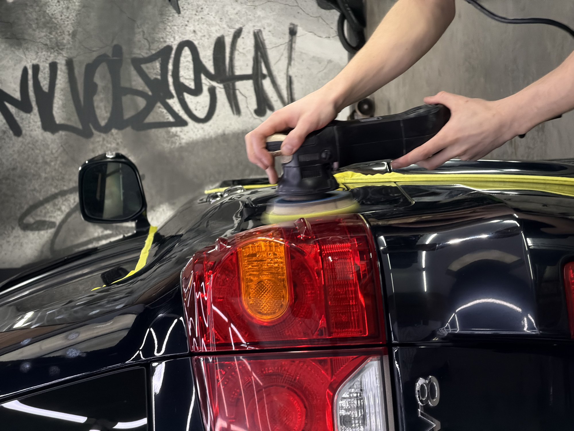 In an indoor setting, a pair of light-skinned human arms are using a black rotary buffer to polish the dark, possibly black, rear quarter panel of a vehicle. Yellow tape is visible along the edges of the body panels. The car's taillight, comprised of red and orange elements, is in the foreground. The vehicle has a "V8" emblem. A side mirror is partially visible on the vehicle's side. The background consists of a concrete wall with grey graffiti. The image does not provide enough information to definitively identify the location as Krasnoyarsk, Russia.
