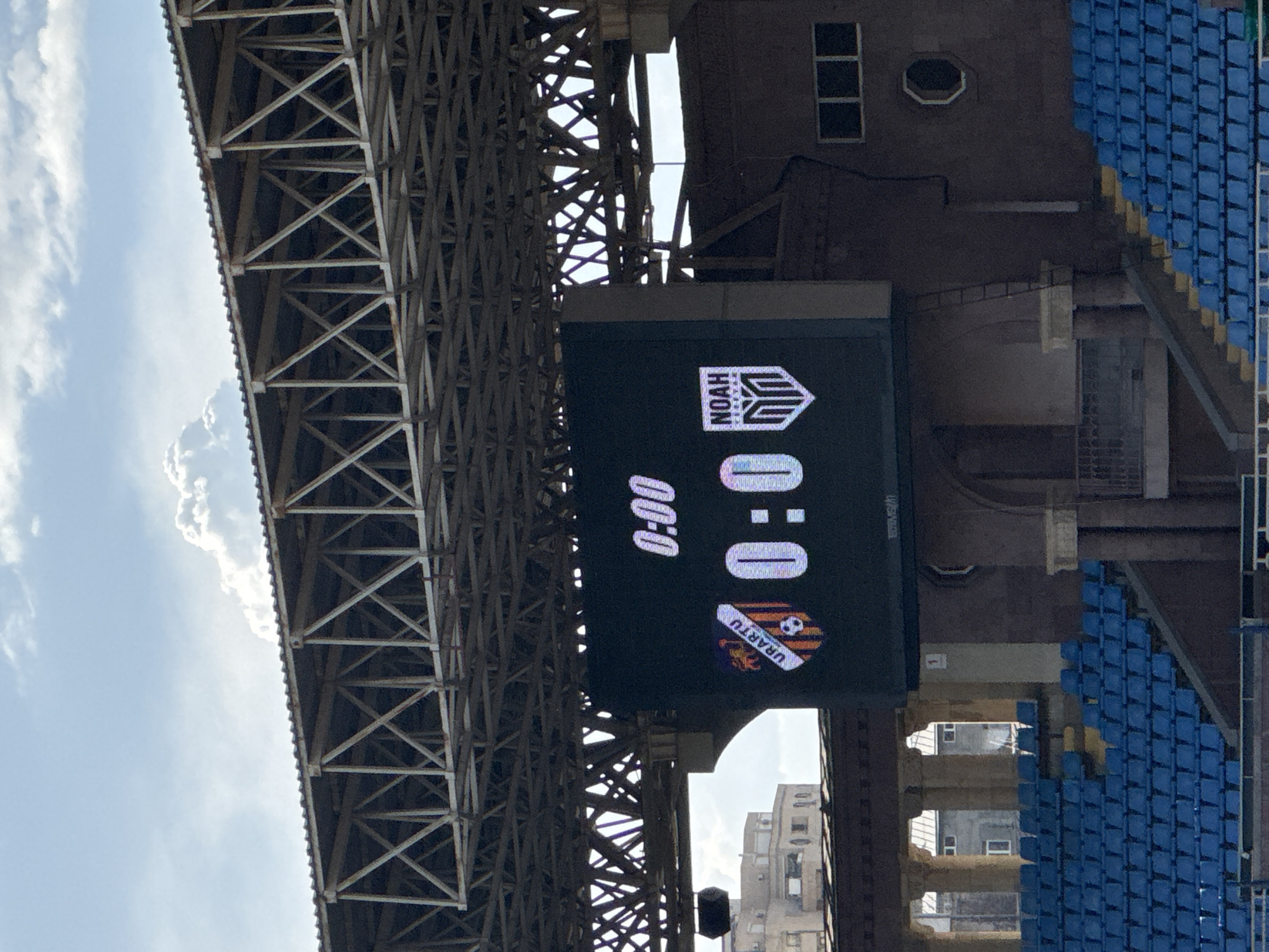 The image depicts a scoreboard at a soccer stadium in Yerevan, Armenia. The scoreboard displays the score as 0:0, with the teams being Urartu and Noah. The stadium has blue seats, and the scoreboard is attached to a brown structure with windows. A steel grid structure is visible above the scoreboard, with the sky and some clouds in the background. Buildings are visible in the distance.