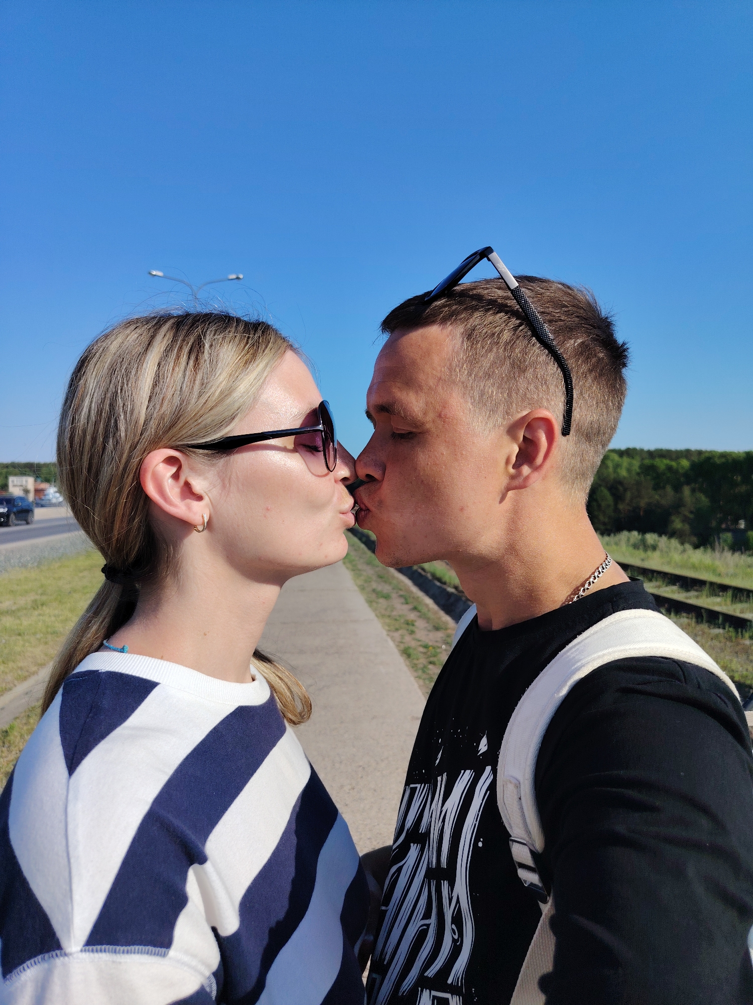 A couple is kissing outdoors on a sunny day. The woman has blonde hair pulled back in a ponytail, sunglasses, and is wearing a striped white and blue shirt. The man has short brown hair with sunglasses on his head and is wearing a black graphic t-shirt and a white backpack. They are standing on a grey sidewalk or path. To the woman's left is a road with cars, and to the man's right is a grassy area with railroad tracks and trees. The sky is clear and blue. The location appears to be in Chaikovsky, Russia, given the road signs and architecture visible in the background. The focus is on the couple, with the background slightly blurred.