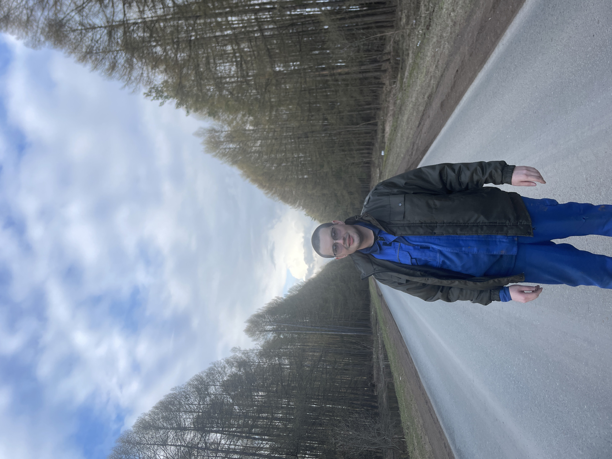 A man is depicted standing on the side of a paved road, looking directly at the camera with a slight smile. He wears glasses, a dark green jacket over a blue long-sleeved shirt, and blue trousers. His hands are visible, one resting by his side and the other slightly raised.
The setting is an outdoor, rural, or semi-rural environment. The road stretches into the distance, flanked on both sides by dense rows of tall, bare trees, indicating a season like late autumn, winter, or early spring. A narrow strip of dirt or grass runs between the road and the tree line. The sky above is partly cloudy, filled with white and grey clouds, suggesting a daytime scene with diffused light. No specific activity beyond posing for the photograph is evident. There is no visible text in the image. Given the location context, this scene is in Podshivalovo, Russia.