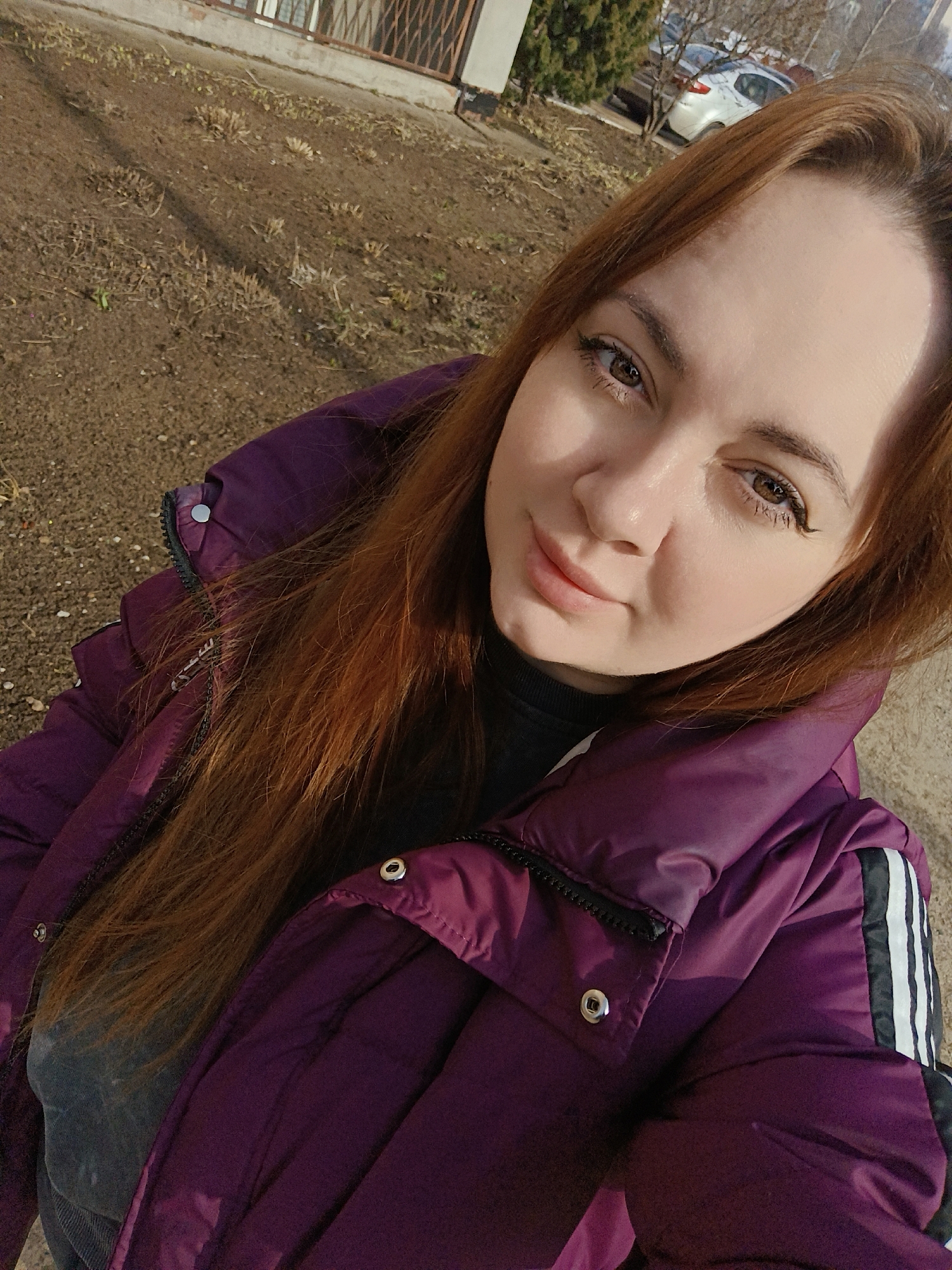 A solo female subject is depicted in a close-up outdoor portrait, likely a selfie, in Chaikovsky, Russia. The woman, with fair skin, brown eyes, and long auburn hair, is looking directly at the camera with a slight smile. She is wearing a dark purple puffer jacket featuring a visible black zipper, silver snap buttons, and a black stripe with three white vertical lines on the upper right sleeve. A dark grey garment is visible beneath the jacket's collar. The immediate background consists of bare, light brown earth with sparse, dry vegetation and small stones. Further back, to the left, a light-colored building with a window covered by a dark metal security grill is visible. To the right, a light-colored sedan car is parked behind bare deciduous trees. The scene is illuminated by natural daylight, casting some shadows on the subject's face.
