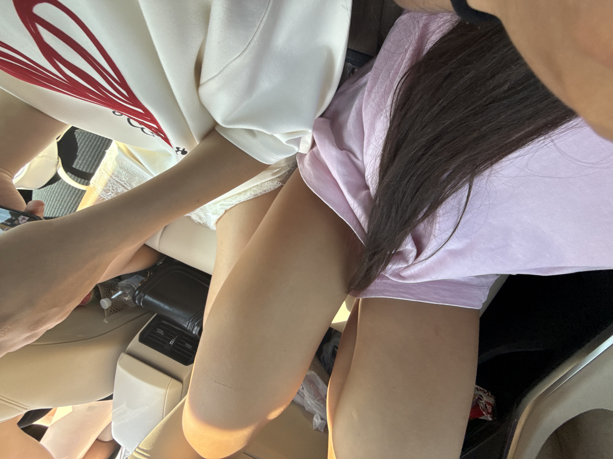 The image depicts an interior view of a vehicle, likely a car, with a focus on the legs and torsos of at least two individuals. The lighting suggests it is daytime, with sunlight illuminating the scene. One person's legs are crossed, and they are wearing a pink garment. Another person's arm, clad in a white sweatshirt with a large red heart design, is visible, and they are holding a smartphone. Between the individuals, a black wallet or purse and a partially visible water bottle in a mesh organizer are present. The vehicle's interior features beige leather or fabric seats. The overall context is an indoor setting within a moving vehicle, suggesting a road trip or journey. No specific landmarks of Arpașu de Jos, Romania, are identifiable within the frame.