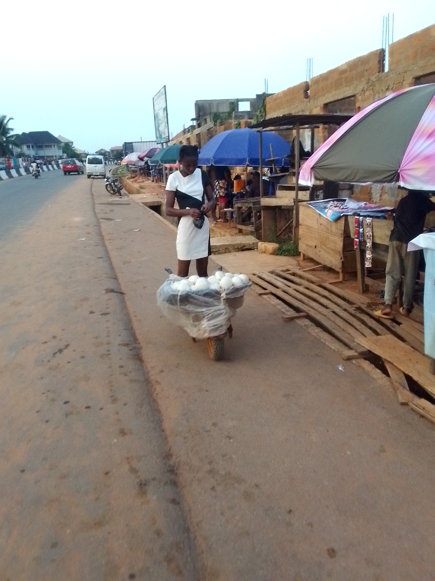 A young woman stands on a street in Orlu, Nigeria, next to a wheelbarrow filled with what appears to be white produce, possibly onions or potatoes. The woman is wearing a white dress with black accents. The wheelbarrow is covered with a clear plastic sheet. Behind her, there are street stalls shaded by umbrellas, with people gathered around them. The street is bordered by a roadside and buildings, including an unfinished brick structure. Several vehicles are visible on the road in the distance.