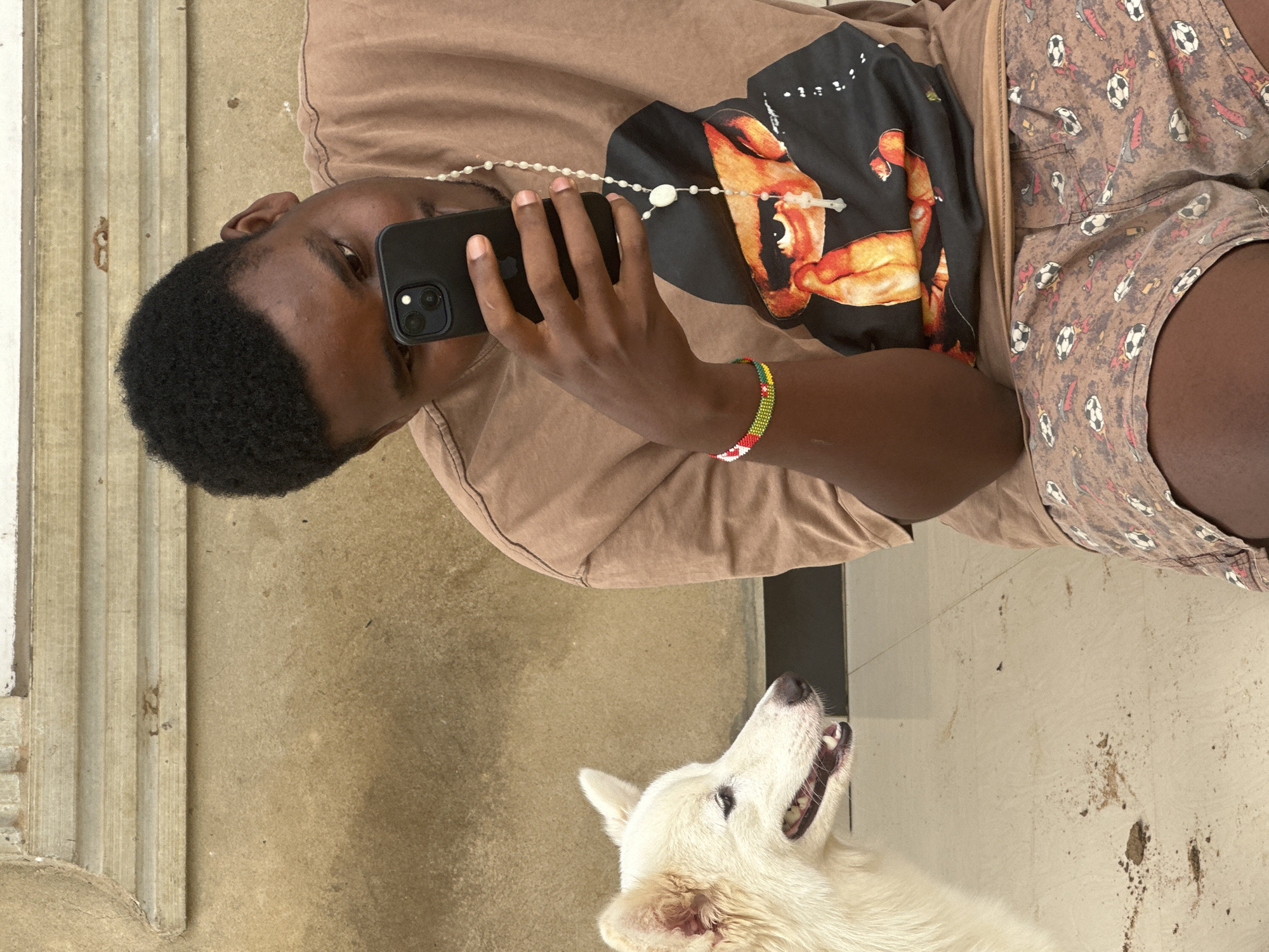 An overhead shot features a young man with short, dark hair using a black smartphone. He is wearing a light brown t-shirt with a graphic print, a beaded necklace, and a colorful beaded bracelet. He is also wearing brown shorts with a soccer ball pattern. Beside him is a white dog with its mouth slightly open and looking upward. The background includes a concrete floor with spots and a part of a fluted wall. The specific location within Ikot Ekpene, Nigeria, cannot be determined without additional information.