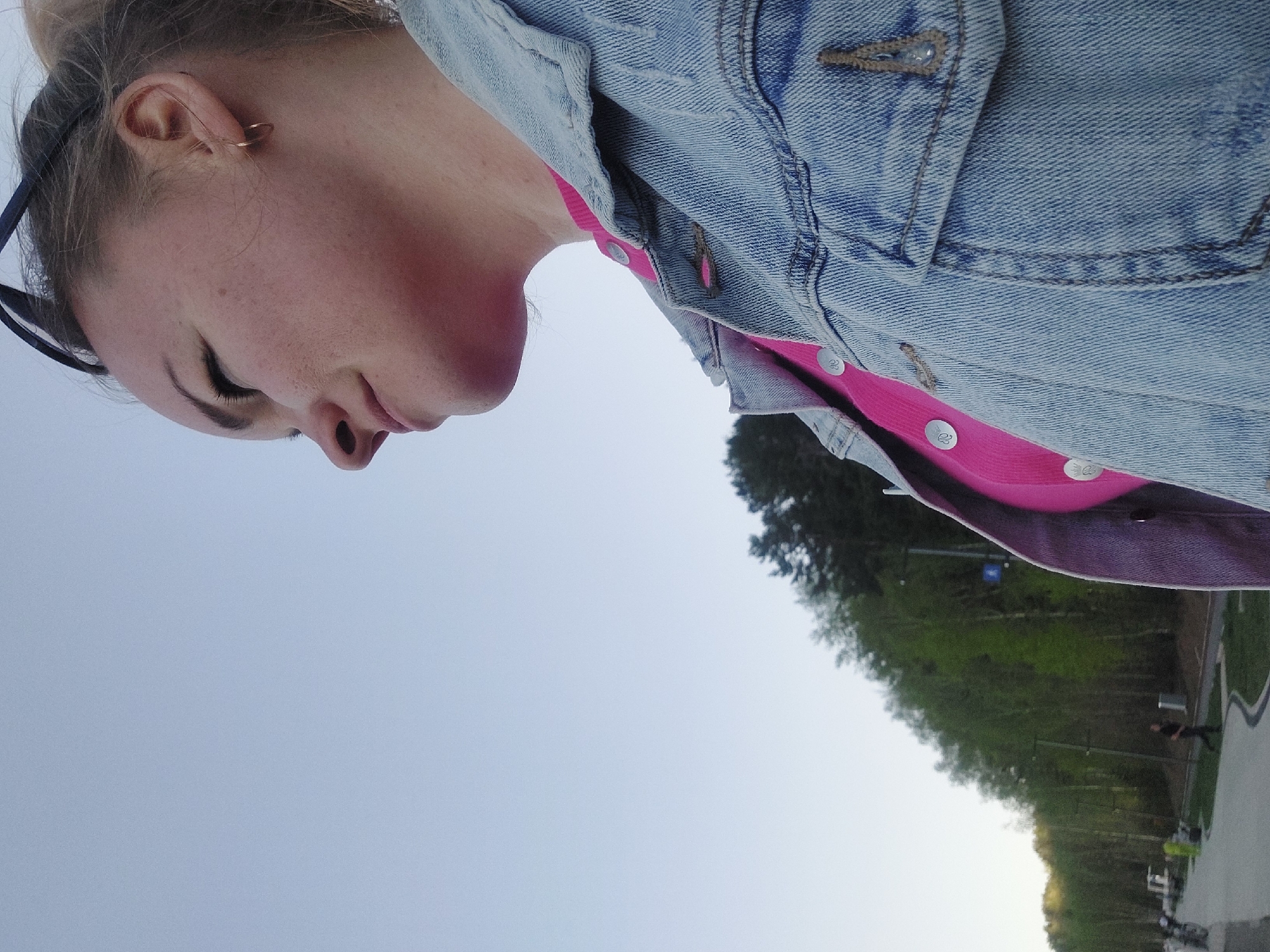 A photograph taken at an oblique angle shows a woman in the foreground, wearing a blue denim jacket over a bright pink shirt with white buttons. Her face, turned to the left, is prominent, displaying closed eyes, freckles, and a gold earring. The background reveals a landscape, likely a park or green space. A line of trees is visible, transitioning into a clear, bright sky. In the lower right, a path or road runs, with a few figures appearing to walk or stand along it. The location is possibly the city of Chaikovsky, Russia, based on implied context.