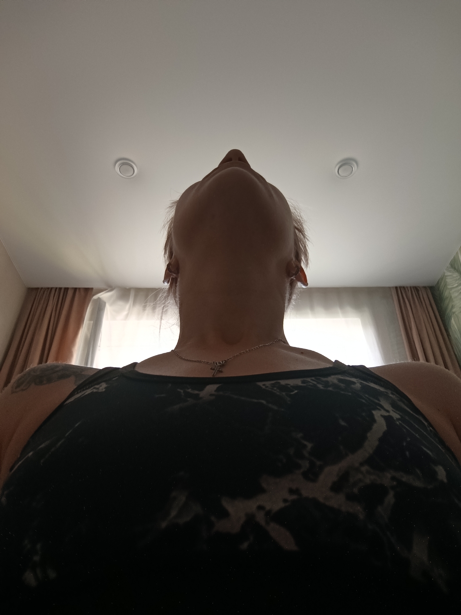 The image is a low-angle shot of a person from the shoulders up, looking upwards towards a ceiling with two recessed lights. The person is wearing a dark tank top with a marbled or tie-dye pattern. A thin silver necklace with a pendant is visible around their neck. The background consists of draped curtains and a window, suggesting an indoor setting. There is a tattoo on the left shoulder. The lighting in the room appears to be natural, coming from the window and artificial light from the ceiling fixtures. The location is an interior space, possibly a bedroom or living area, in Novokuybyshevsk, Russia.
