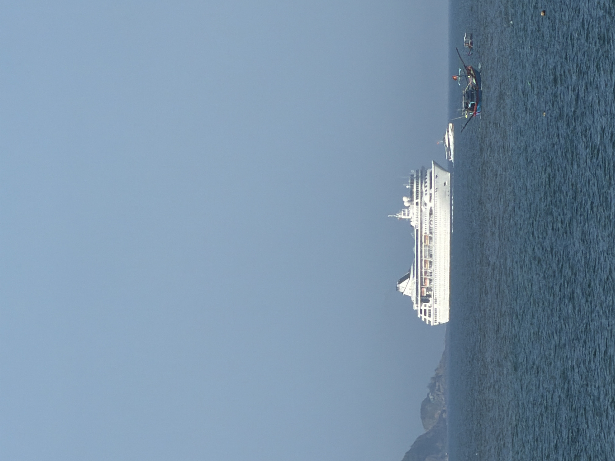 The image depicts a panoramic view of the sea off the coast of Nha Trang, Vietnam. In the distance, a large white cruise ship is sailing on the calm, deep blue water under a clear, light blue sky. To the left of the cruise ship, a small, rocky island with sparse vegetation is visible, partially obscured by haze. Closer to the foreground, several smaller boats are present. One is a traditional Vietnamese fishing boat with a dark blue hull and a red flag, its crew appears to be tending to equipment. To the right of the fishing boat, another small boat is anchored. The water exhibits gentle ripples, suggesting a tranquil maritime environment. The scene is characteristic of the coastline of Nha Trang, known for its scenic beaches and numerous islands.