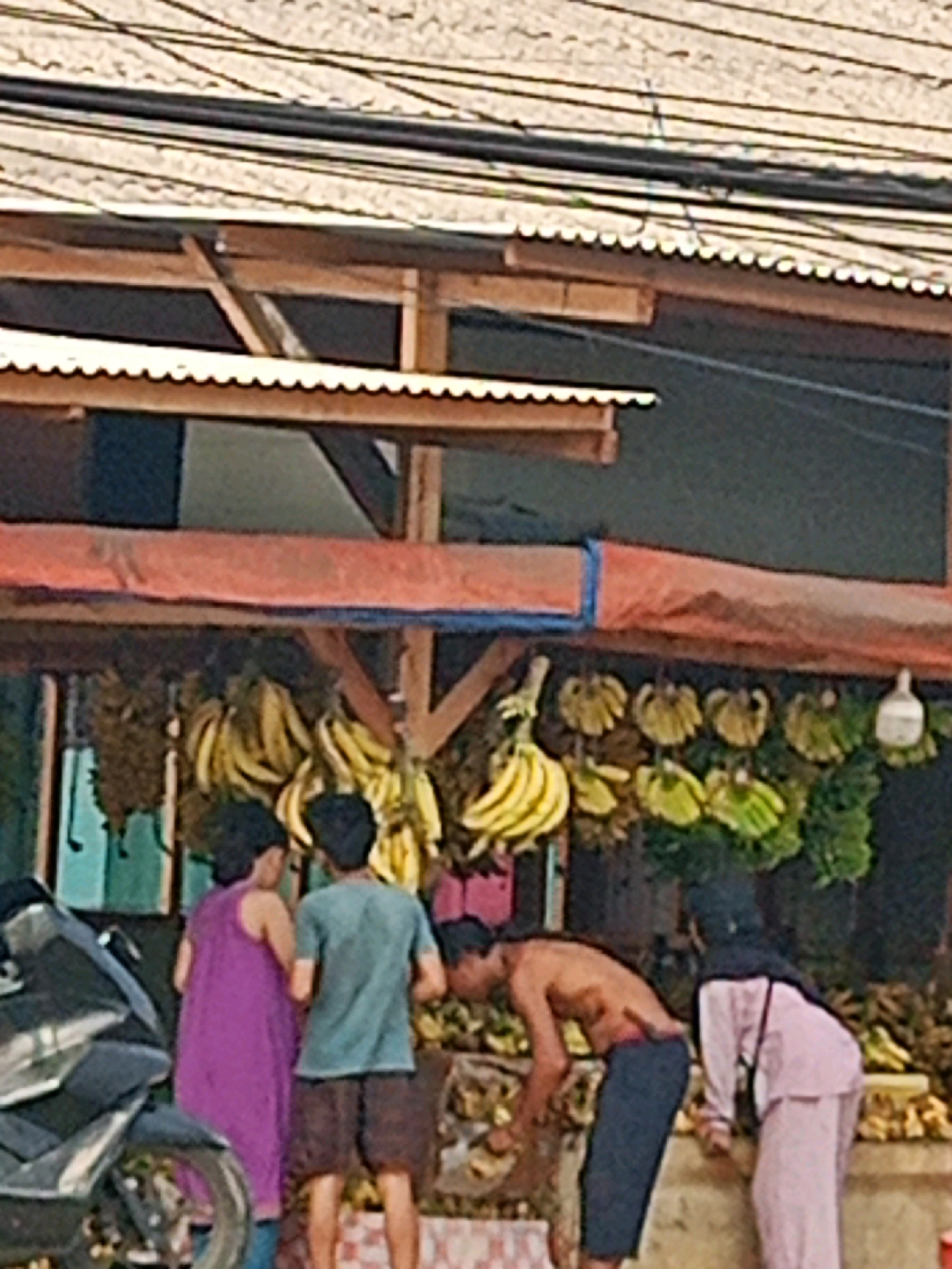 The image depicts an outdoor market scene, likely in Sriamur, Indonesia, under a corrugated metal roof supported by wooden beams. A variety of fruits, predominantly bunches of bananas, are displayed on a table in the foreground. Two women, one in a purple dress and the other in a pink headscarf and outfit, are standing near the fruit stall. A man, shirtless and wearing dark shorts, is bent over, seemingly interacting with the produce. Another man, wearing a teal t-shirt and brown shorts, stands next to the woman in the purple dress. A black motorcycle is parked to the left of the frame. The background shows a stall structure with a reddish awning. Electrical wires are visible above the market canopy.