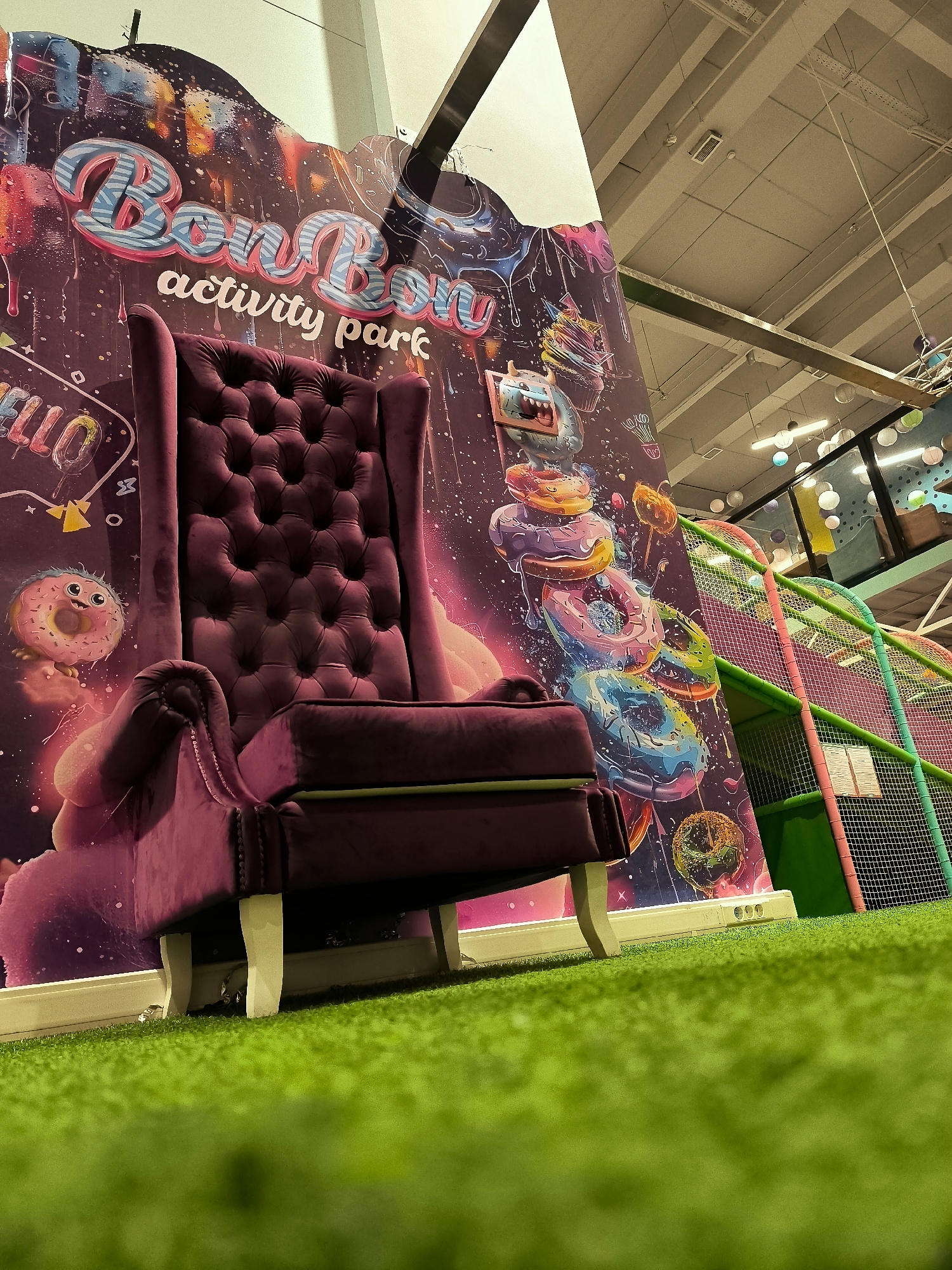 In a brightly lit indoor space with a green, artificial turf floor, a plush, dark purple velvet armchair with a high back and tufted upholstery sits prominently. The armchair is positioned in front of a vibrantly decorated wall mural. The mural features the words "BonBon activity park" in stylized lettering, accompanied by whimsical illustrations of donuts, a cartoon monster, and various colorful, abstract designs with a cosmic theme. To the right of the armchair and mural, a section of a multi-colored indoor playground structure is visible, characterized by green and pink netting and tubes. The ceiling above shows exposed industrial elements like beams and wiring, along with circular light fixtures and some ventilation equipment. The overall scene suggests a children's entertainment venue. The location is identified as Qarağandı city administration, Kazakhstan.