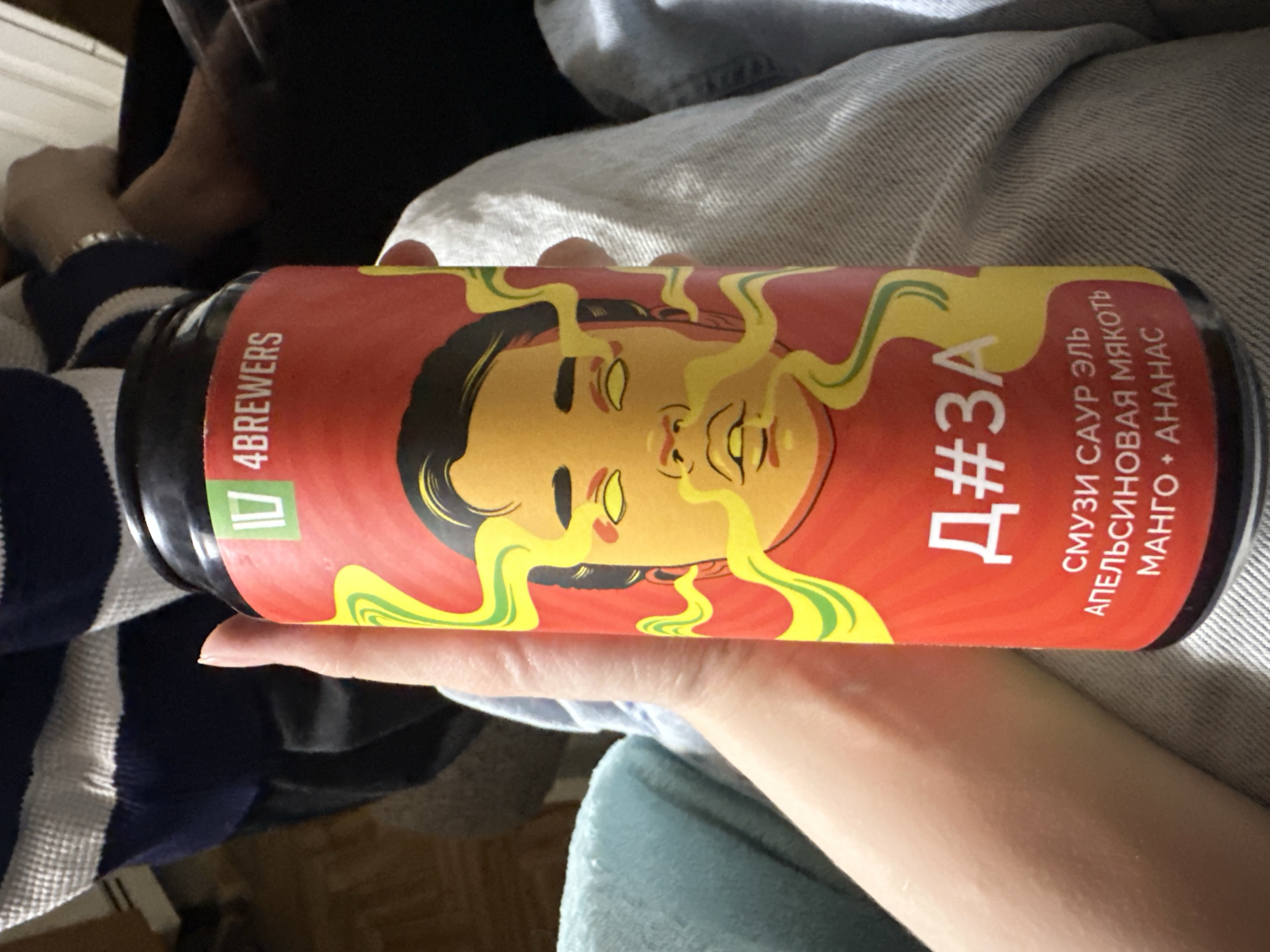 A person holds a can of 4Brewers beer, specifically a "Smoothie Sour Ale" with orange, mango, and pineapple flavors, written in Russian. The can's design features a yellow illustration of a person's face with radiating lines, set against a red background. The surrounding environment includes clothing, suggesting an indoor setting. There is no visual evidence to confirm the location in Moscow, Russia, though the Russian text on the can implies a possible connection.
