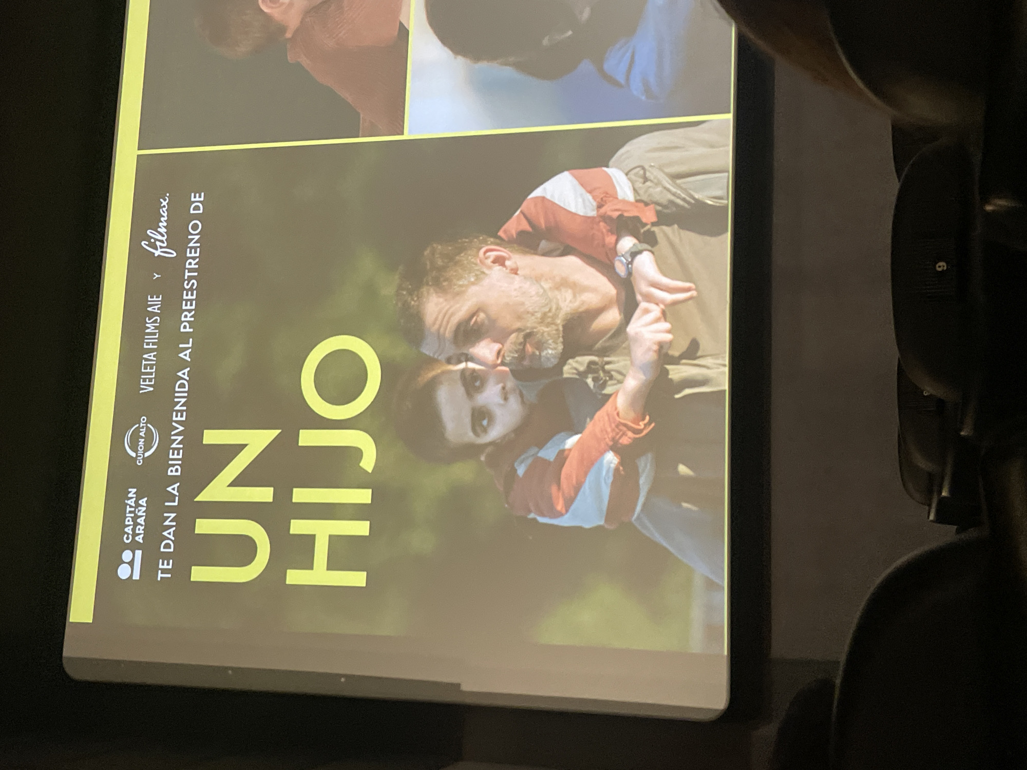 The image shows a movie poster for "Un Hijo," a film by Capitan Arana. The poster is positioned in a theater setting, with theater seats visible in the foreground. The poster also features the logos of Guion Alto, Veleta Films AIE, and Filmax. The poster contains a photograph of a bearded man hugging a younger person, likely a child, with an outdoor, slightly blurred background. The film's title, "UN HIJO," is printed in large, bold yellow letters. The tagline "TE DAN LA BIENVENIDA AL PREESTRENO DE" is printed above the title. The picture does not allow for a location within the city of Barcelona to be definitively assessed.