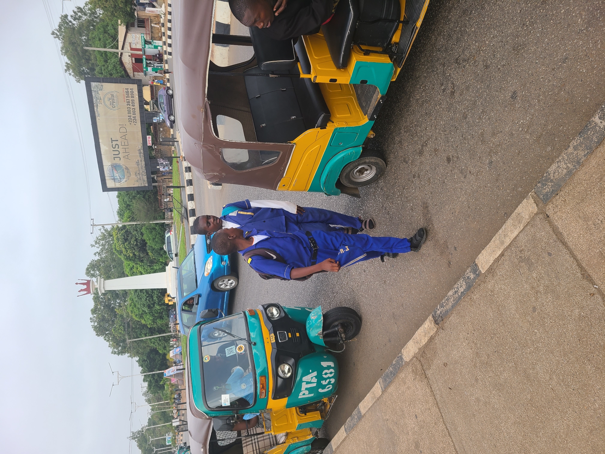 The image shows a street scene in Jos, Nigeria, with three-wheeled vehicles known as "keke" or "tricycles" prominently featured. Two men wearing blue uniforms are standing in the street next to one of the tricycles. There is a billboard advertising "Crystal" visible in the background. Cars and other vehicles are also present, indicating traffic. A monument with a crown on top can be seen in the background. The tricycle in the foreground has "PTA-6581" written on it.