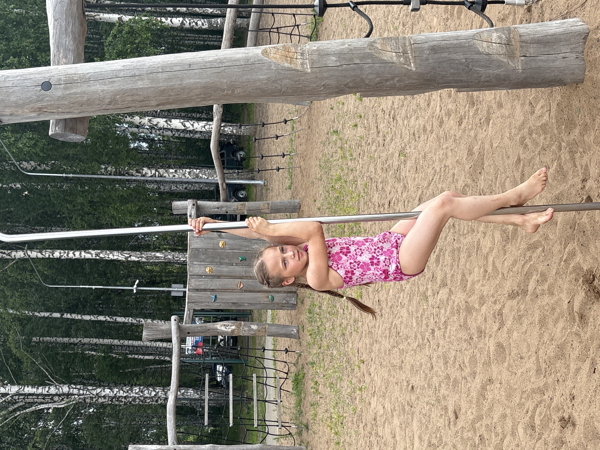 A young girl with blonde hair in a pink floral swimsuit is hanging from a horizontal metal bar at a playground. She is upside down, with her legs bent at the knees and her arms extended, gripping the bar. Her face is turned towards the camera, and she has a slight smile. Her hair is braided and hangs down. The playground has a sandy ground and wooden structures. In the background, there are many tall birch trees with white bark and green foliage. The sky is not visible, but the lighting suggests it is daytime. The overall mood is playful and active. A rope climbing net and wooden platforms are visible on the left side of the image. The image is oriented vertically.