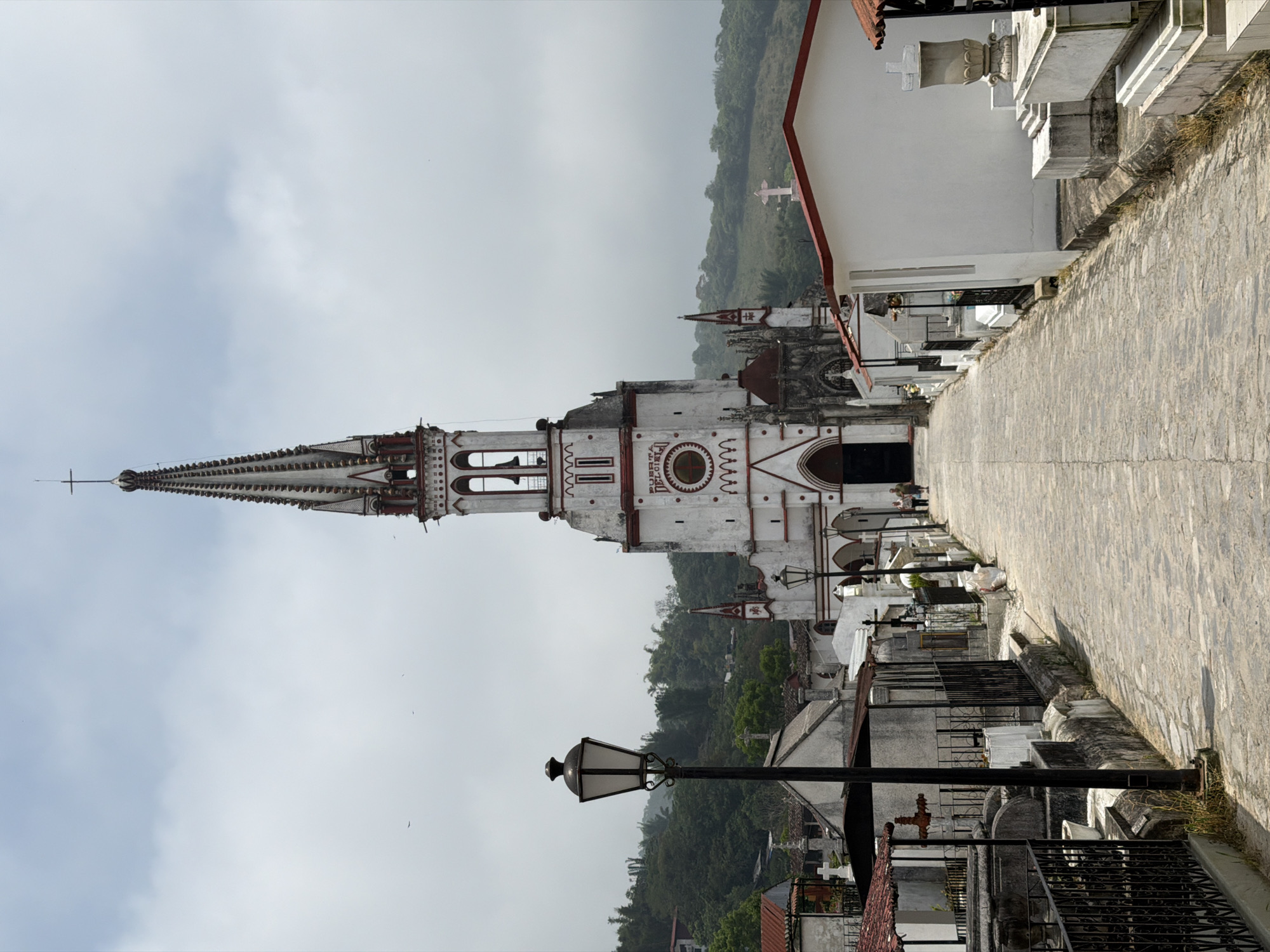 The image shows a white and red-trimmed church with a tall, thin spire, in Cuetzalan, Mexico. The church is positioned adjacent to a cobblestone path that appears to run through a cemetery, indicated by numerous tombstones and crosses. A lamppost is visible in the lower part of the frame, standing amidst structures and gravesites. In the background, a hill or mountain is seen, partially obscured by a hazy sky.