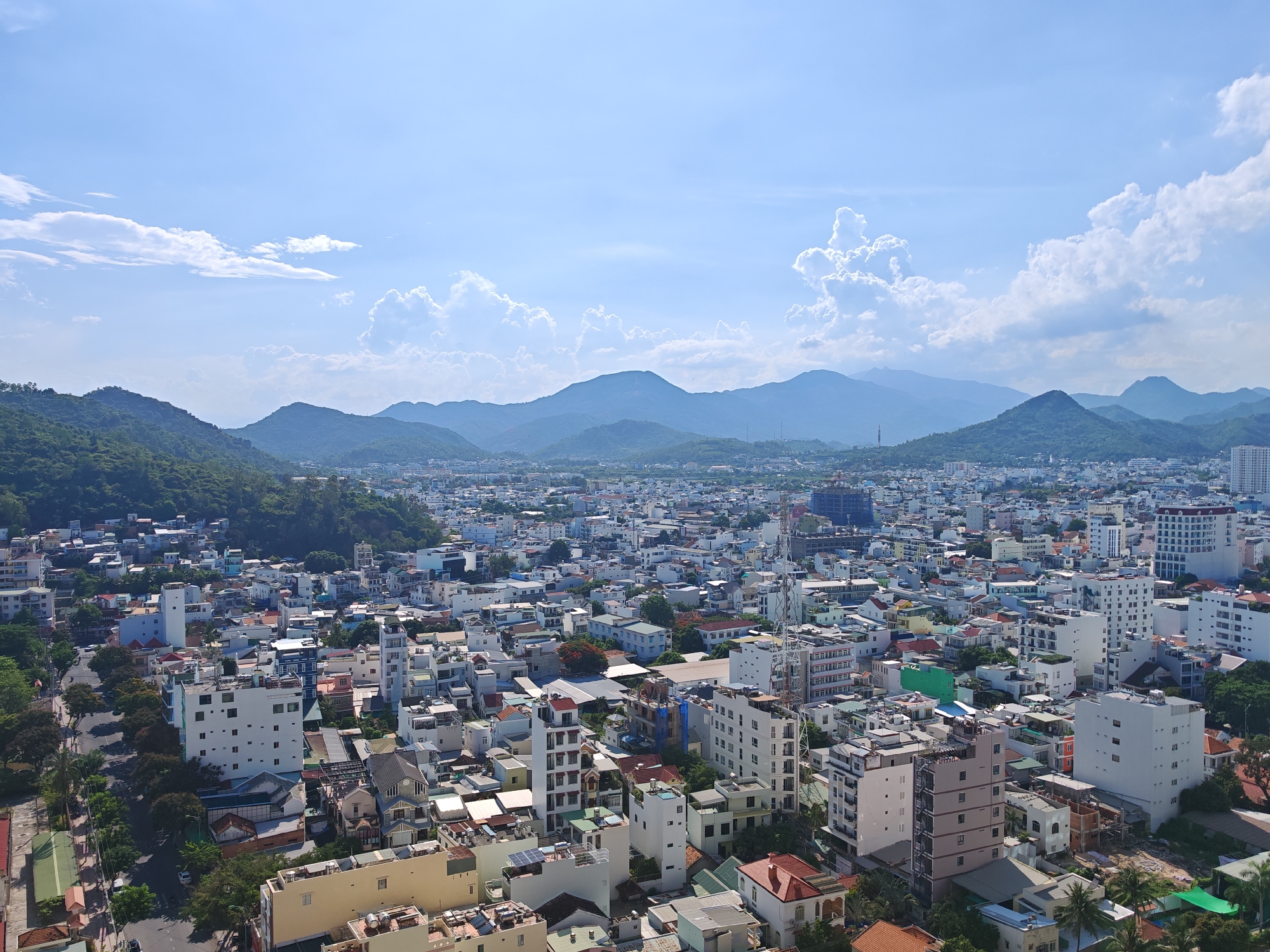 An aerial, wide-angle, high-resolution photograph captures a sprawling urban landscape in Bac Nha Trang Ward, Vietnam, under a bright, partly cloudy blue sky. The city extends outwards, a dense patchwork of residential and commercial buildings in various heights and architectural styles, predominantly white, cream, and pastel colors, with occasional splashes of green, blue, and red rooftops. Streets, some lined with trees, snake through the dense urban fabric, with a few cars visible. In the foreground, the closest buildings are modern, multi-story structures, some with balconies and visible air conditioning units. Further into the city, the buildings become more varied, with a mix of apartment blocks, smaller houses, and some industrial-looking structures. In the distance, a range of lush green mountains forms a dramatic backdrop to the city, their slopes covered in dense vegetation. The sky is dotted with fluffy white clouds, creating a sense of depth and atmosphere. The overall impression is one of a vibrant, densely populated coastal city nestled against a mountainous terrain.