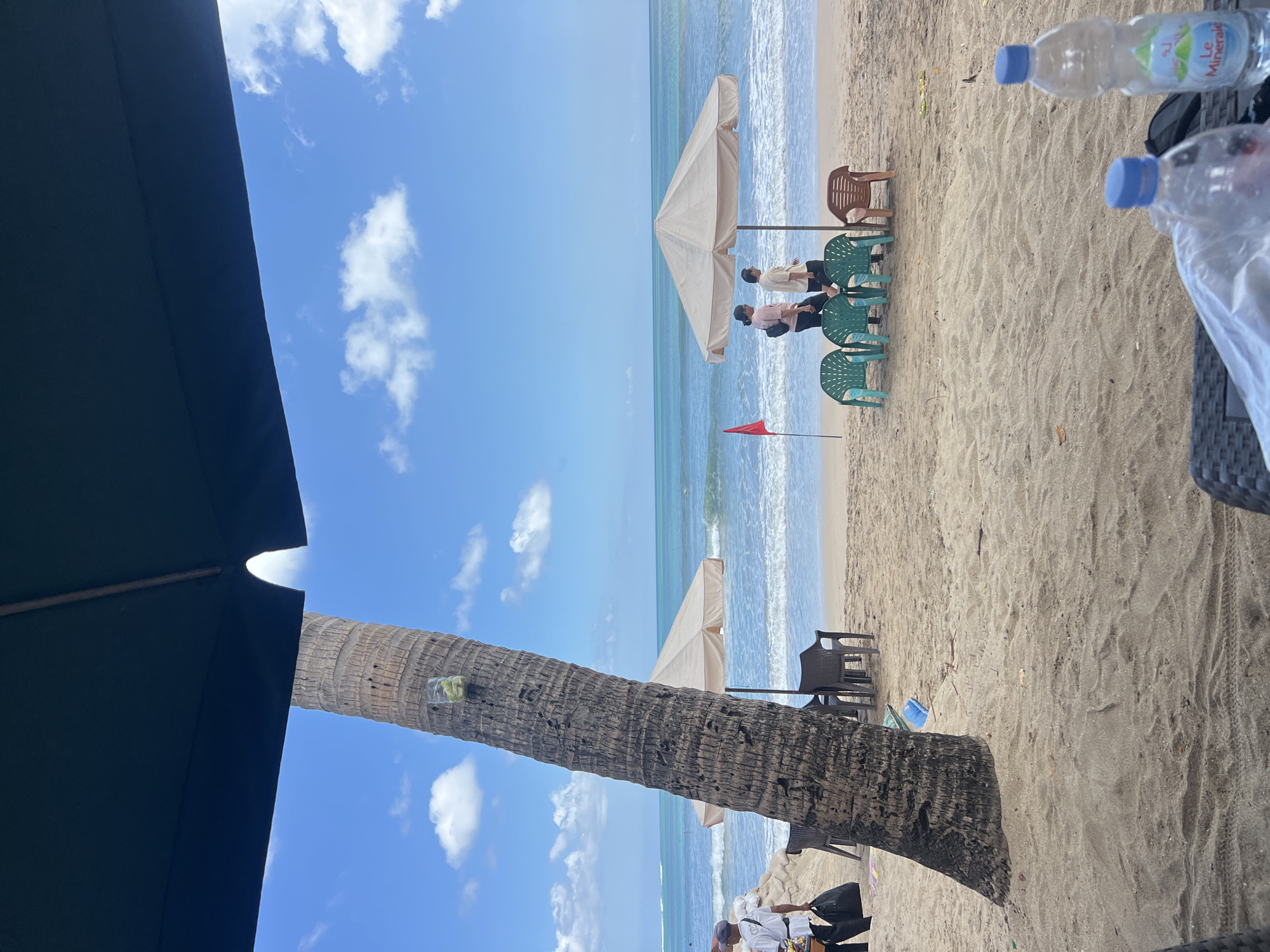 The image depicts a beach scene in Kuta, Indonesia, under a bright blue sky with scattered white clouds. In the foreground, a dark umbrella casts a shadow, partially obscuring a portion of the frame. A prominent palm tree stands to the left, with a glass containing a drink attached to its trunk.
Further back, the sandy beach stretches towards the ocean. Two beige beach umbrellas are set up, providing shade. Several plastic chairs are arranged around them, some in green and one in brown. Two individuals are walking near one of the umbrellas, facing away from the camera. A red flag is planted in the sand to the left of the umbrellas.
The ocean is calm with gentle waves rolling onto the shore. In the distance, a few people can be seen in the water. To the far left, a person in a white shirt and dark pants is walking with a black bag. In the foreground, on a woven surface, two plastic water bottles are visible, one with a blue cap and the other with a white cap and a label that reads "Le Mineral". The overall atmosphere suggests a relaxed day at the beach.