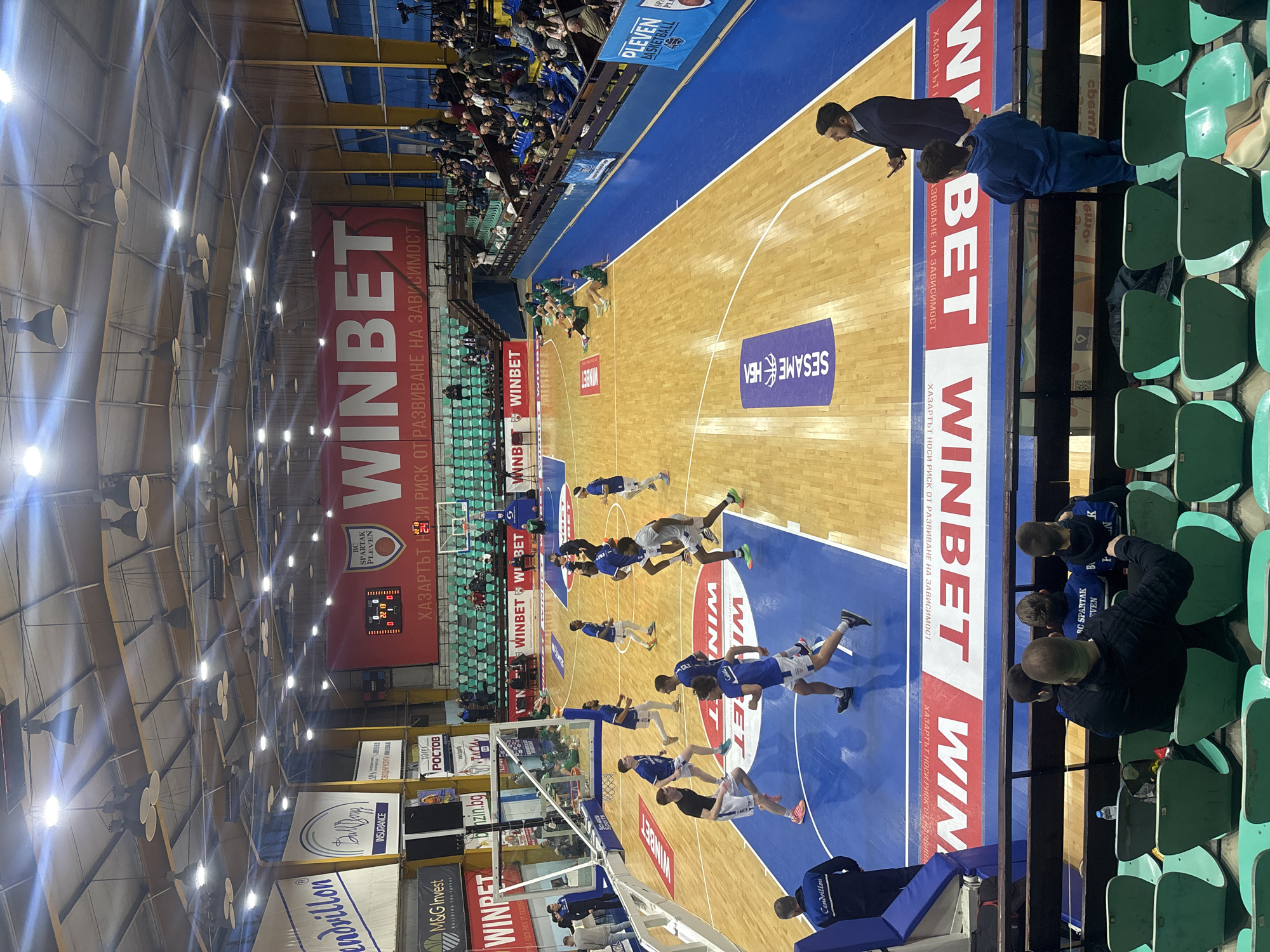 This image depicts an indoor basketball game in progress within an arena located in Pleven, Bulgaria, as evidenced by banners displaying "BC SPARTAK PLEVEN" and "PLEVEN BASKETBALL." The scene is captured from an elevated position in the spectator stands.
The basketball court features a light brown wooden surface with blue painted areas for the key and sidelines. White lines delineate court boundaries and the three-point arc. Several players are visible on the court; one team is primarily dressed in blue jerseys with white shorts, while at least one player wears a grey jersey with white shorts. A group of players in green tracksuits are seated along the far sideline. On the near side of the court, a coach or team staff member in a dark tracksuit with "Kendrillon" on the back stands near the baseline. Another man in a dark suit holds a phone while standing by the sideline.
The stands, composed of tiered seating in shades of green and blue, are moderately filled with spectators. In the immediate foreground, a few individuals, including an adult male with three children, are seated, observing the game.
The arena ceiling is high, featuring numerous suspended lighting fixtures illuminating the court. Large banners hang from the arena walls, predominantly displaying sponsorships and team information. A prominent red banner with white lettering reads "WINBET" and includes Bulgarian text below. Another large banner on the opposite side identifies "BC SPARTAK PLEVEN" alongside a scoreboard displaying "22:13" as the time and scores of "0" for both home and guest teams, indicating an early stage or warm-up. Additional sponsor banners in Bulgarian are visible throughout the venue. A professional camera on a tripod is positioned in the upper right stands, suggesting media coverage.
