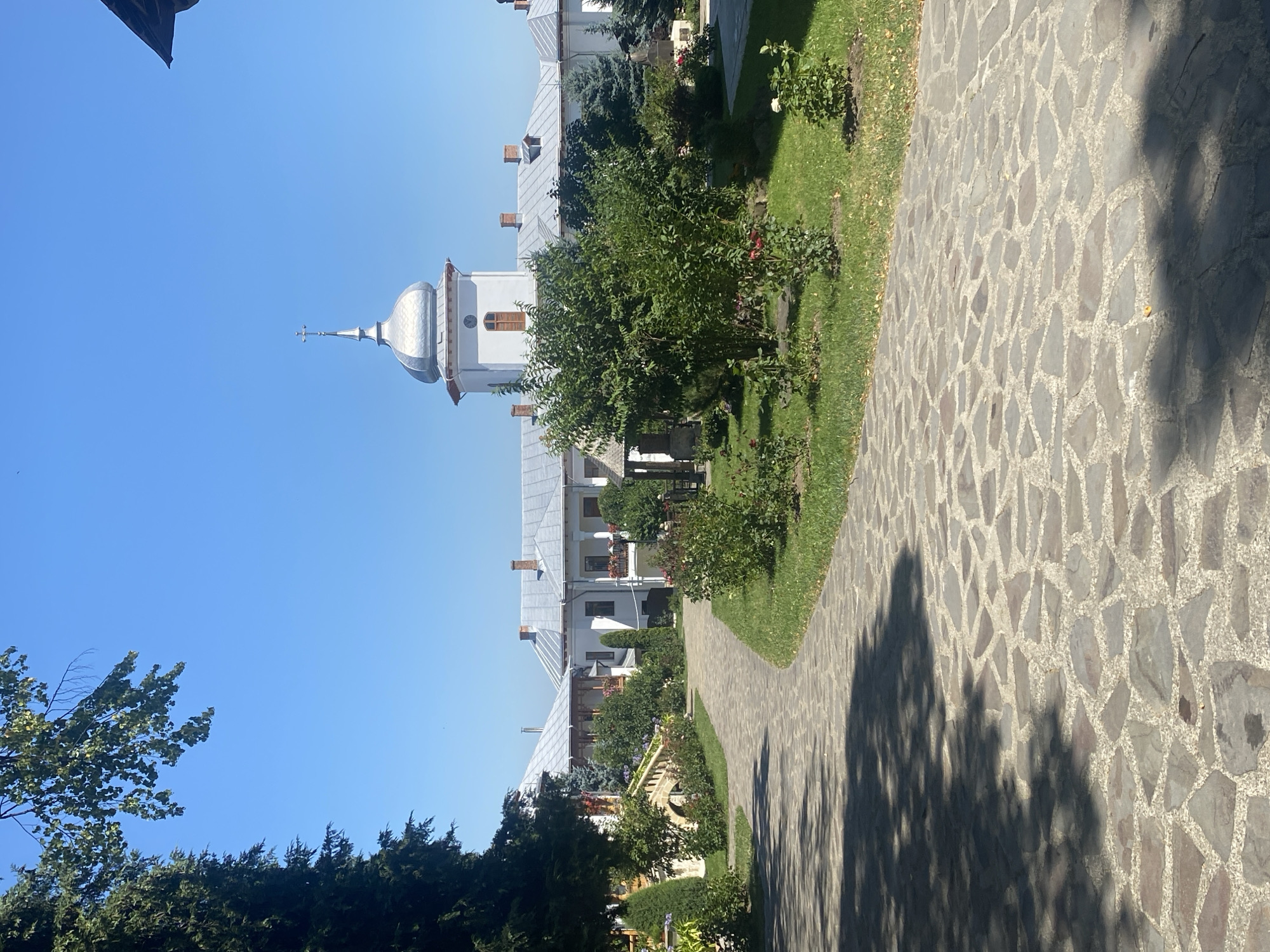 The image depicts a serene monastery courtyard in Filioara, Romania, on a clear, sunny day. A paved pathway, constructed from irregular grey and brown stones, curves through the foreground, partially shaded by the dappled sunlight filtering through overhead tree branches. Flanking the pathway are meticulously maintained green lawns, dotted with various shrubs and rose bushes, some of which are in bloom with red and white flowers.
In the middle ground, the monastery buildings present a traditional architectural style. They are primarily white, with pitched grey roofs and dark red brick chimneys. One prominent structure features a silver, onion-shaped dome topped with a cross, adding a distinctive religious element to the scene. Several windows are visible, some with decorative wooden shutters and flower boxes. A stone bridge with railings also arches over a section of the pathway.
The background is dominated by a bright blue sky. Evergreen trees and deciduous trees with lush green foliage frame the scene, adding to the tranquil atmosphere of the monastery grounds. No people are visible in the image, contributing to the sense of peace and solitude.