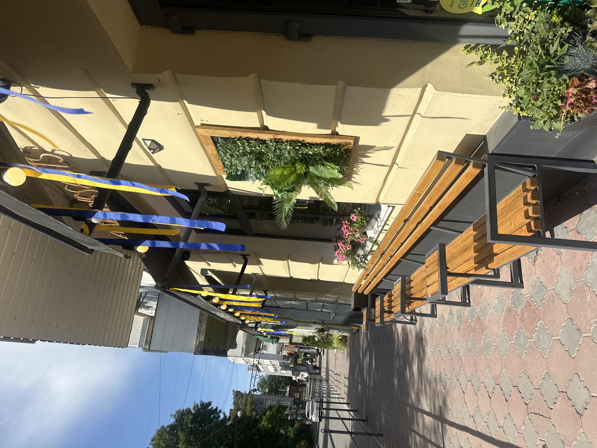 A sidewalk cafe with wooden benches and yellow and blue ribbons decorating the building. The cafe is located in Ternopil, Ukraine, and is situated on a street with trees and buildings in the background. There are a few people walking on the sidewalk in the distance. The weather appears to be sunny.