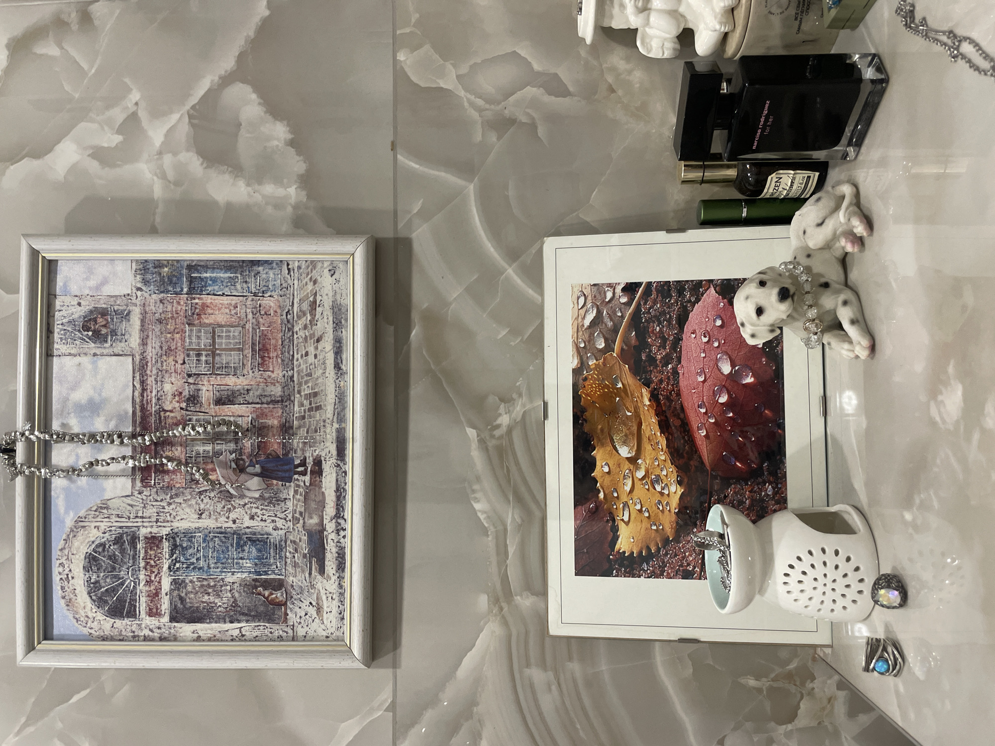The image depicts a section of a wall, possibly in a home in Krasnoyarsk, Russia. The wall has a marble-like pattern. On the left side of the image, there's a framed artwork of a building with an archway, and a silver necklace is draped over it. To the right, there's another framed image depicting leaves with water droplets. Near the second picture, there's a small porcelain dog figurine, a white ceramic object with holes, and a few pieces of jewelry. Additionally, there's a perfume bottle and other cosmetic products on top of the wall section.