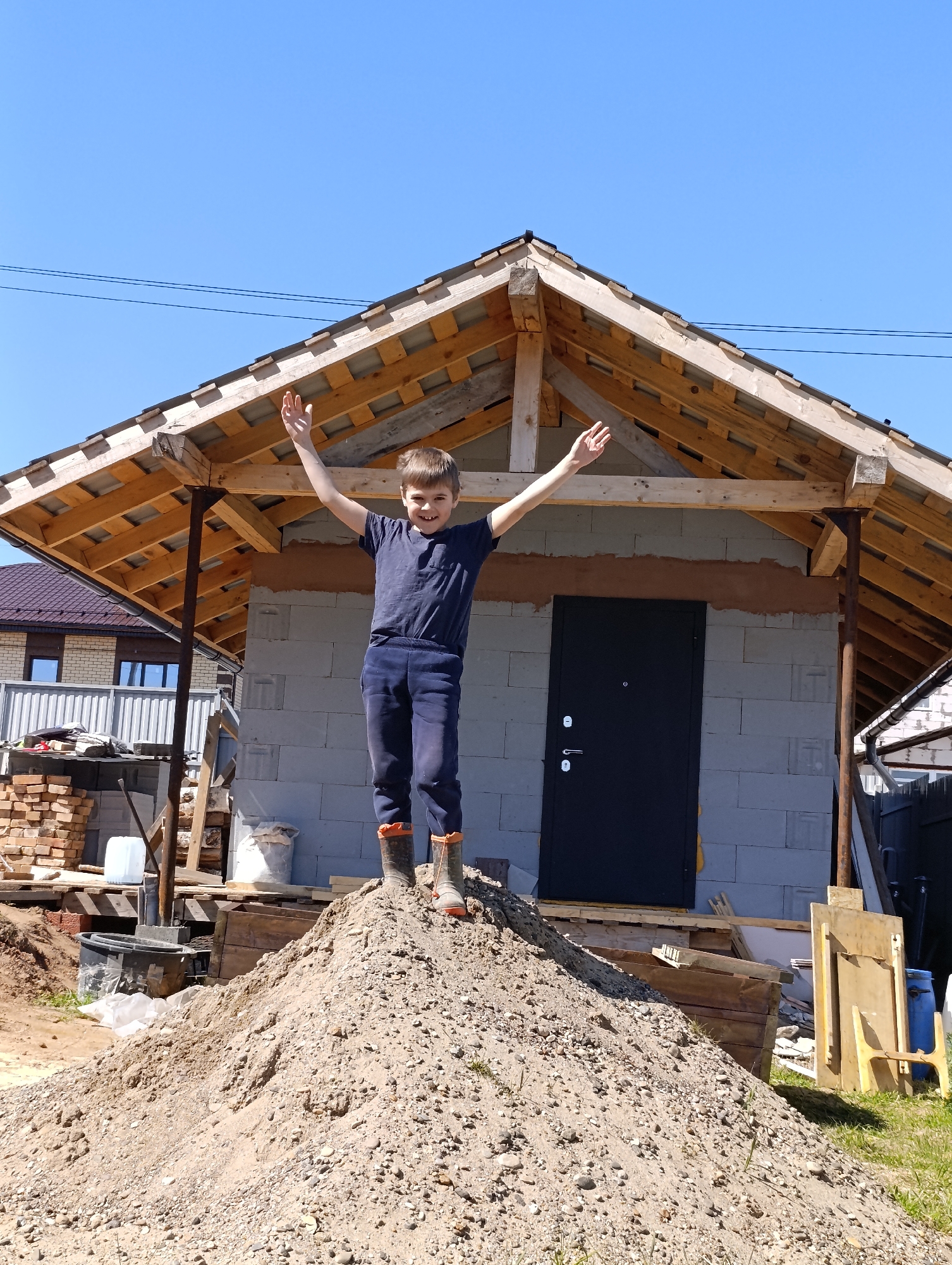 The image depicts a young boy standing atop a large pile of sand or dirt, with arms raised in a celebratory gesture. He is wearing dark clothing and boots. Behind him is a partially constructed building with a visible wooden roof frame and block walls with a dark door. The background includes a clear blue sky and glimpses of other buildings and construction materials, suggesting a construction site or residential area under development. The location is Chaikovsky, Russia.