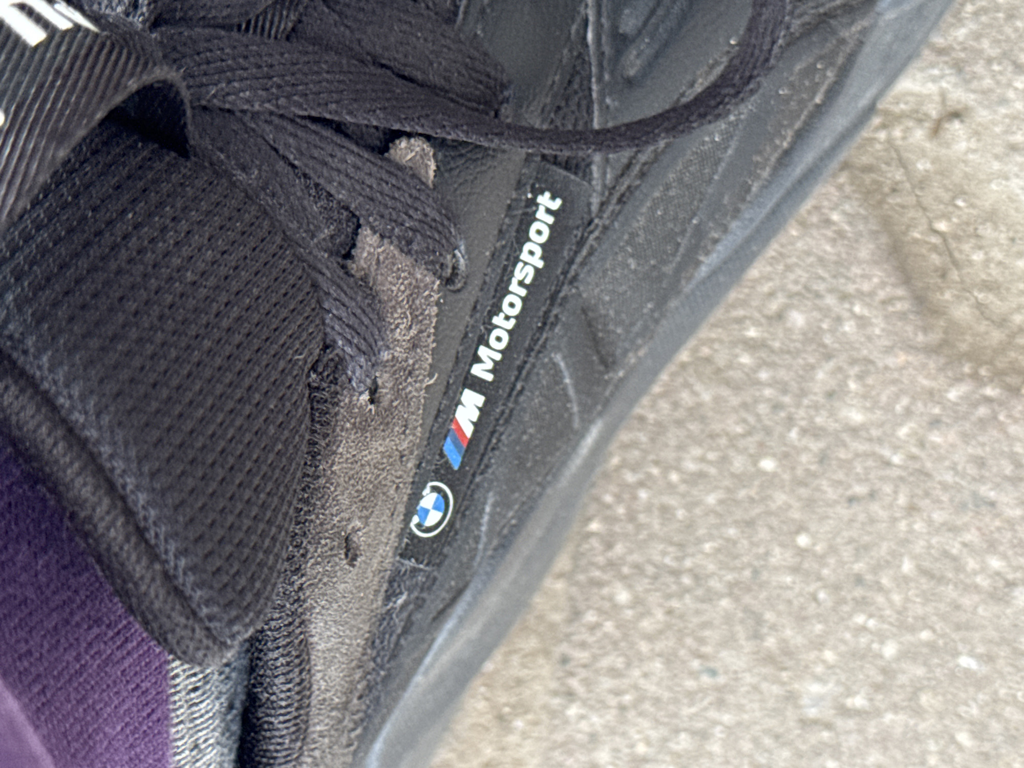 The image shows a close-up of a black sneaker with black laces. The side of the shoe features the BMW Motorsport logo and branding. The shoe appears to be worn and slightly dirty. The background consists of a concrete surface. No information suggests this is in Plovdiv, Bulgaria.