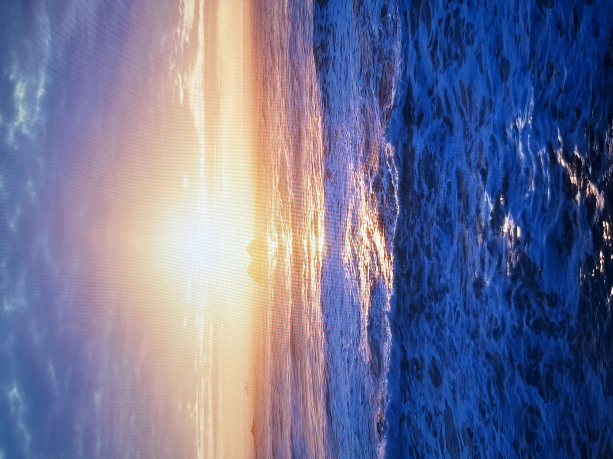 The image depicts a seascape at sunset. The sun, a bright orb, is positioned low on the horizon, casting a golden and pink glow across the sky and the ocean's surface. The sky is a gradient of colors, transitioning from deep blue at the top to soft purples, oranges, and yellows near the horizon. Wispy clouds are scattered across the sky, catching the sunlight. The ocean is active, with visible waves and white foam. A reflection of the setting sun shimmers on the water. In the distance, two small islands or rock formations are visible, silhouetted against the bright sky. The foreground of the ocean is a vibrant, deep blue, with the crests of waves illuminated by the fading light. The overall scene conveys a sense of natural beauty and tranquility. This image is not located in Kyiv, Ukraine, as it depicts a natural seascape with islands, which are not characteristic of Kyiv's urban landscape.