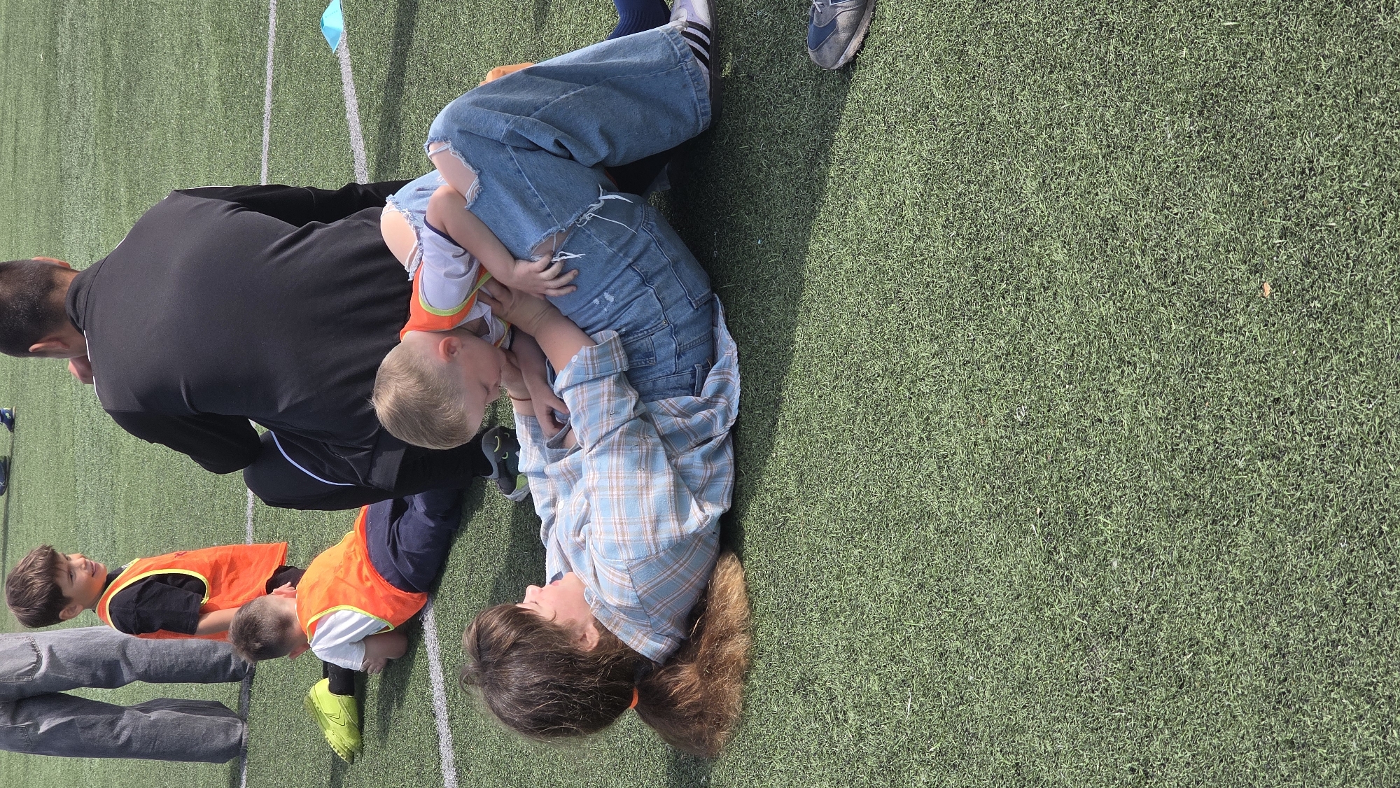 On a grassy field in Unknown, Unknown, a woman lies on her back, cradling a young child in her arms. The woman has long brown hair and wears a blue plaid shirt and ripped blue jeans. The child wears an orange and white shirt. A man in a black jacket kneels nearby, and two other children in orange vests are also present.