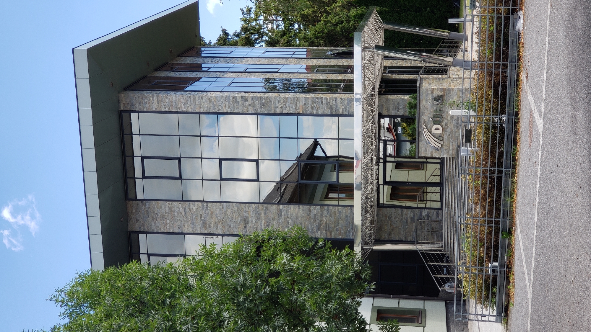 A modern, multi-story building with a stone and glass facade stands under a blue sky with scattered clouds. A large, leafy green tree is positioned to the left of the building, partially obscuring the view. The building's entrance features a covered walkway with a metal, lattice-like ceiling, leading to a set of stairs and double glass doors. The facade is composed of alternating sections of light-colored stone and large, reflective glass panels, which mirror the sky and surrounding environment. The word "DÚHA" is visible on a stone plaque near the entrance. A metal fence encloses a landscaped area with bushes in front of the building, and a paved area, likely a parking lot, is in the foreground. The overall scene depicts an office building in an urban or suburban setting. This is the Dúha, a.s. headquarters in Prešov, Slovakia.