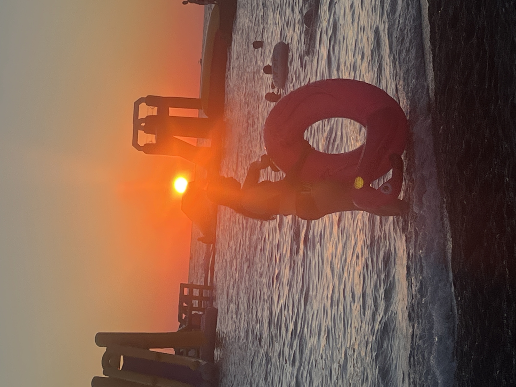 The image depicts a sunset over a beach, likely in Sochi, Russia, given the context. In the foreground, a child is standing in the shallow water, holding a large red inflatable ring. The child is partially silhouetted against the setting sun, which is a bright orange orb low in the sky, casting a warm glow over the scene. The water is calm with small ripples, and waves are gently breaking on the shore. In the background, several inflatable water slides and structures are visible, suggesting a water park or recreational area. Other people can be seen in the water, some with inflatable rings, enjoying the evening. The overall atmosphere is serene and tranquil.