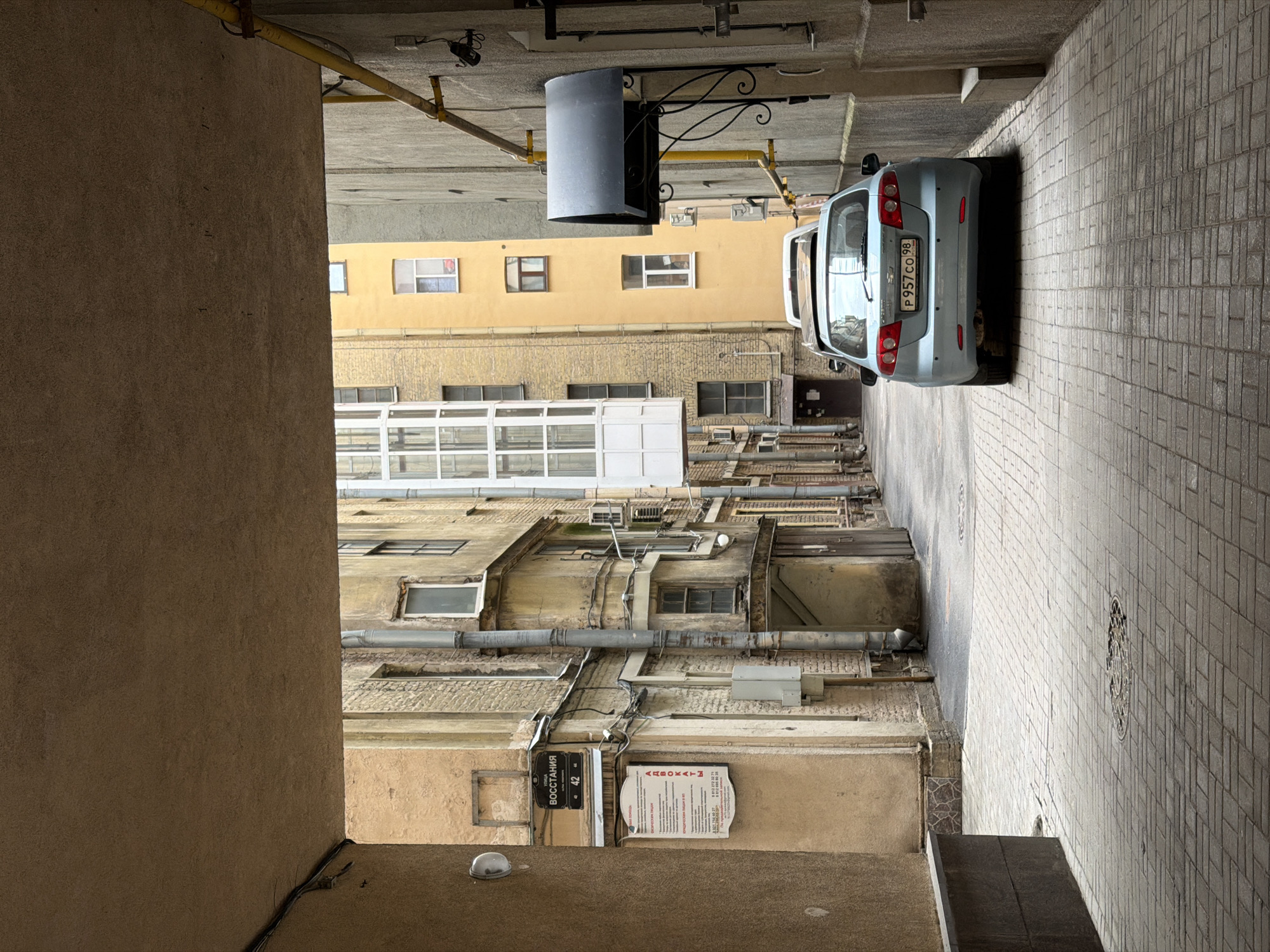 A slightly rotated, medium, and elevated-angle shot from within a St. Petersburg, Russia courtyard shows two sides of the courtyard with an automobile between them. The courtyard sides are adjacent to each other. The shot is taken from an enclosed location on the left, from which the courtyard is viewed.
On the left side is a textured wall of tan-colored material. On the opposite side of the alleyway, across from the vantage point, is a sign stating “ВОССТАНИЯ 42”. A commercial sign for “АВОКАТЫ” is to the right of that. At the bottom of the shot, a light grey helmet sits on the ground near the wall. Further into the shot and above the signs, building materials and architecture are apparent. In the middle of the shot and higher up, piping runs the length of the buildings. Multiple windows of varying sizes and styles are observed along the walls of the courtyard.
In the upper right quadrant of the shot, an automobile is seen at a slight angle along the right wall. The license plate on the car reads “P957CO98”. The right side of the courtyard is paved with light grey cobblestones. Pipes and mechanical apparatus run along the top of the shot. A grey hanging light fixture is mounted above the car.