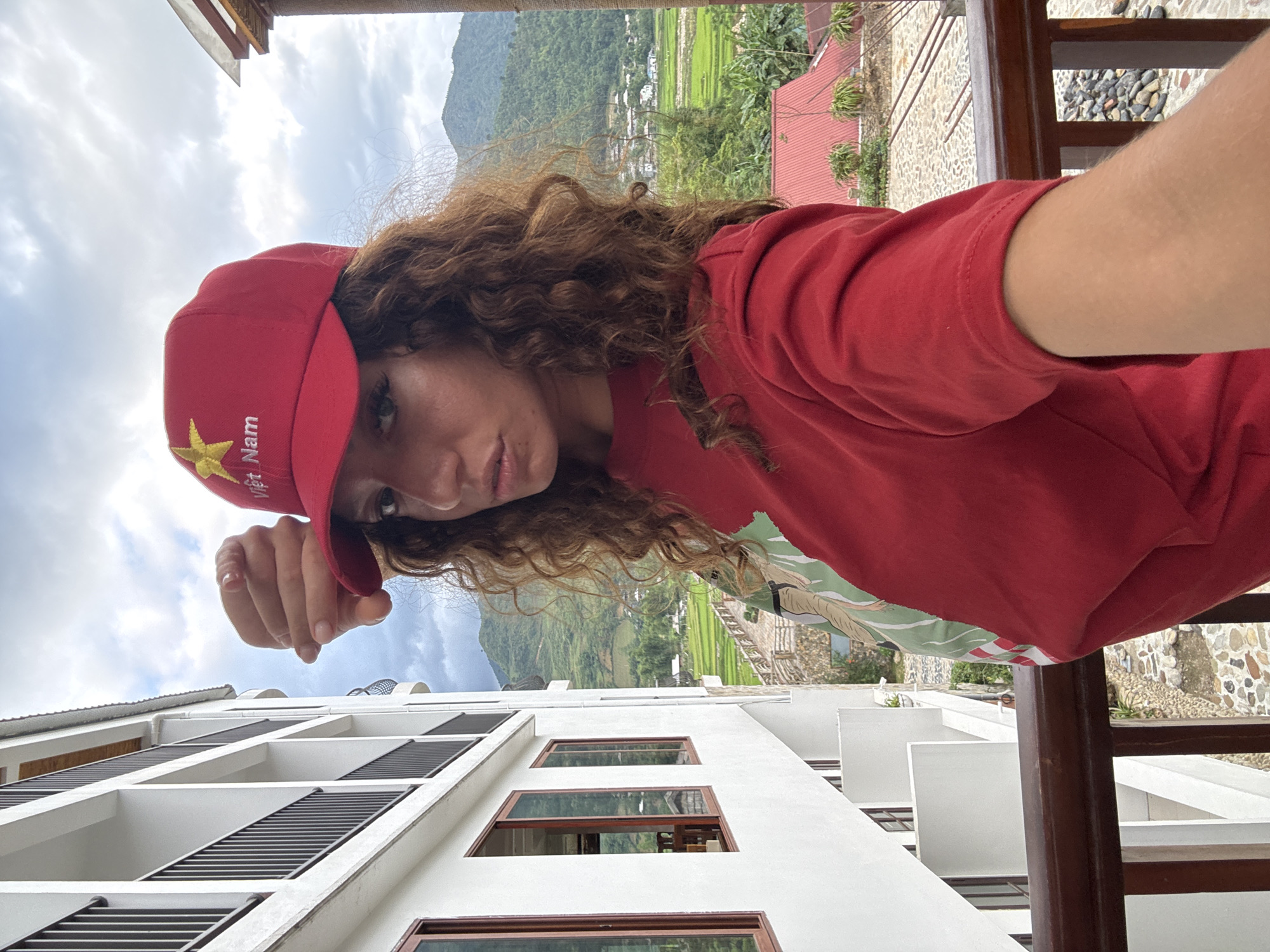 A woman with curly brown hair and wearing a red baseball cap with a gold star and the word "Việt Nam" on it is the focal point of this selfie-style image. She is also wearing a red t-shirt and a ring on her left hand. The shot is taken from an elevated perspective, with a white, multi-story building featuring dark-framed windows visible in the foreground below her.
In the background, a landscape of green rice paddies and rolling hills is visible, suggesting a rural or semi-rural environment. To the right, a wooden railing is present. A stone paved area is visible.
The location is likely in the city of Tú Lệ, Vietnam, based on the text on the baseball cap.
