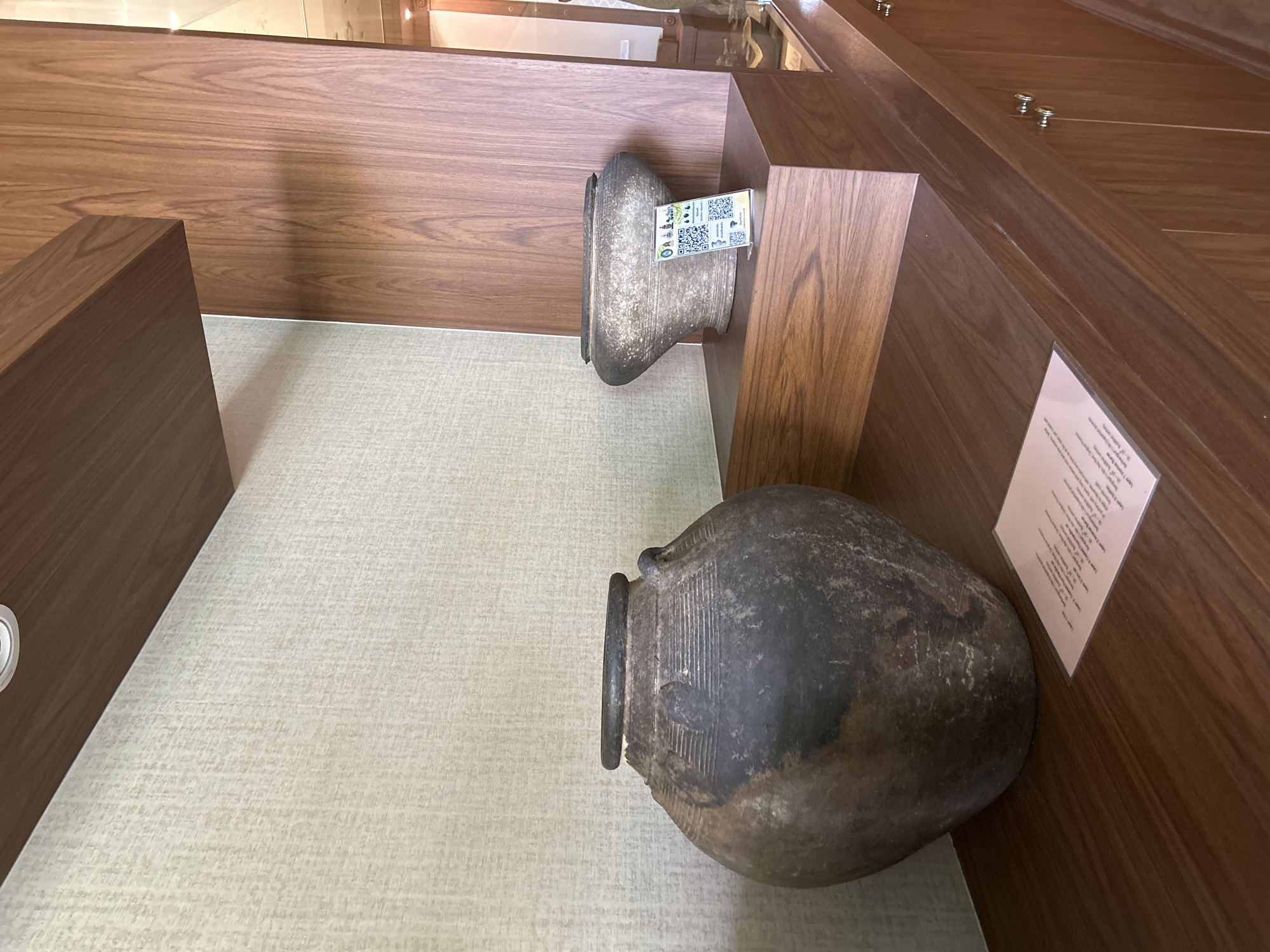 A close-up, eye-level shot displays two ancient pottery vessels on a wooden shelf. The larger, dark-colored vessel sits on the left, with a dark rim and two handles. A small, light pink card with text is placed in front of it. To the right, a lighter, grayish-brown vessel rests on a wooden block, featuring a QR code display. The background consists of a textured, light-colored wall on the left and a wooden panel on the right. The image was taken in the Thai Ko Subdistrict, Thailand.