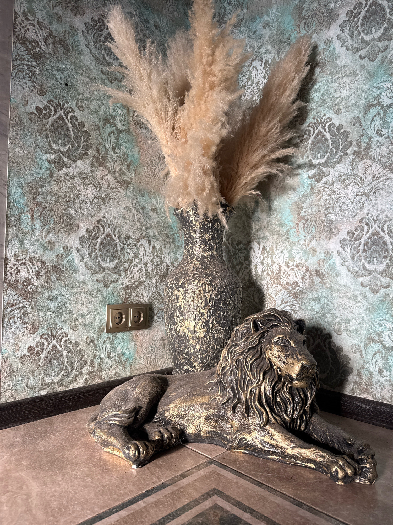 This is a photograph of a corner of a room with decorative wallpaper and a tiled floor. A large, ornate vase filled with dried pampas grass stands against the wallpaper. In front of the vase, a decorative lion statue is lying down. There is an electrical outlet on the wall. The lighting suggests an indoor setting.