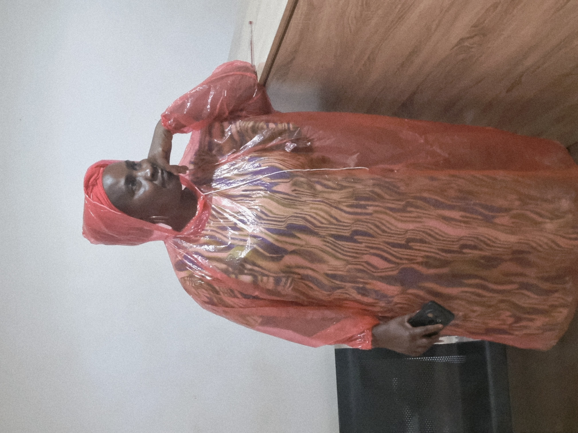 A woman, appearing to be of African descent, is standing indoors against a light-colored wall. She is wearing a hooded, translucent pink poncho over a patterned garment with wavy, vertical stripes of purple, gold, and brown. The poncho has a slight sheen and appears to be made of plastic. She has a silver hoop earring in her left ear. Her right hand is resting on her chin, and her gaze is directed to her right, away from the camera. Her left hand is holding a dark-colored smartphone. To the right of the woman is a wooden surface, possibly a counter or desk, with a light wood grain. To her left, a dark, textured surface, possibly a chair or part of a bench, is visible. The lighting suggests an indoor environment, possibly a waiting area or a public space. The image quality is grainy. The provided context places the scene in Jos, Nigeria.